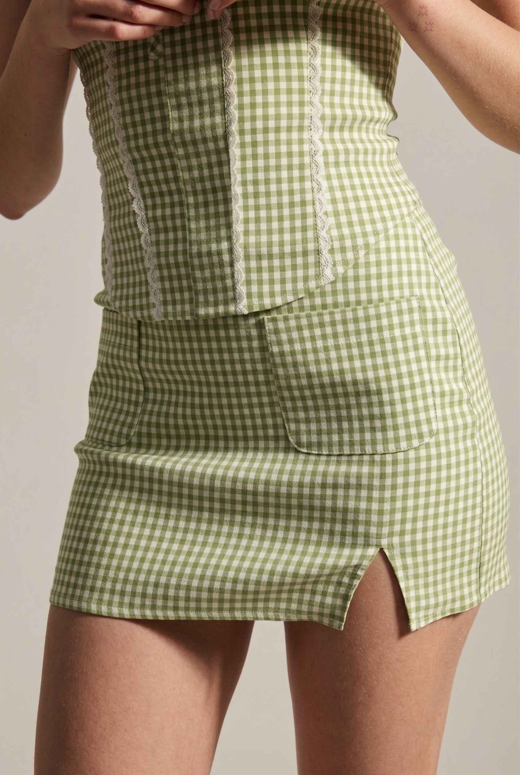 Model wearing Poppy Skirt in Sage Green Gingham Mini Skirt with Pockets paired matching corset tank top close up slit detail