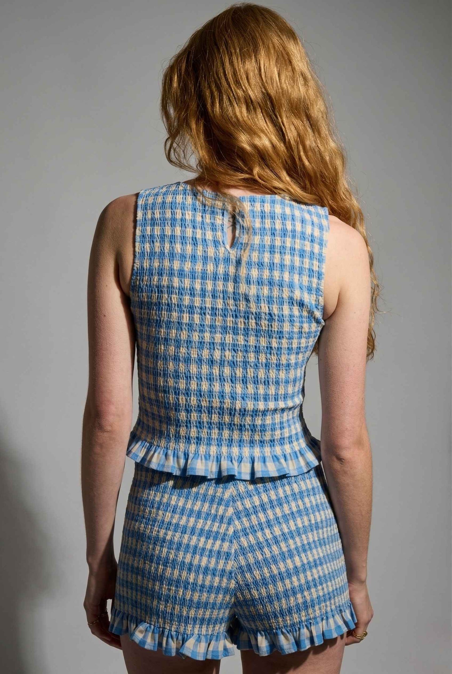Person wearing a Poppy Top Blue Gingham Smocked Sleeveless Knit with a peplum top and shorts on a gray background