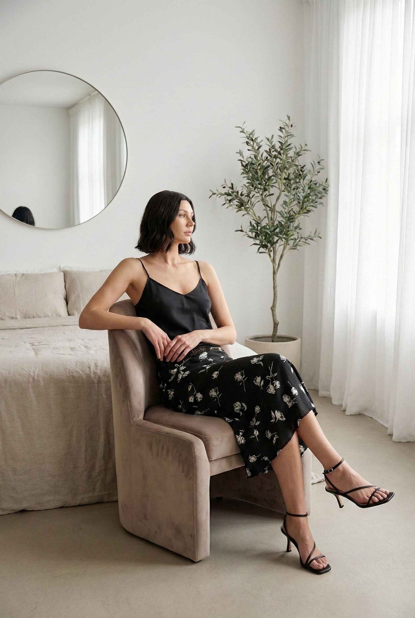 Model wearing Raven Skirt in Black Floral Satin Midi Skirt with Lace Waist paired with silk camisole and strappy heels posing in minimalist bedroom
