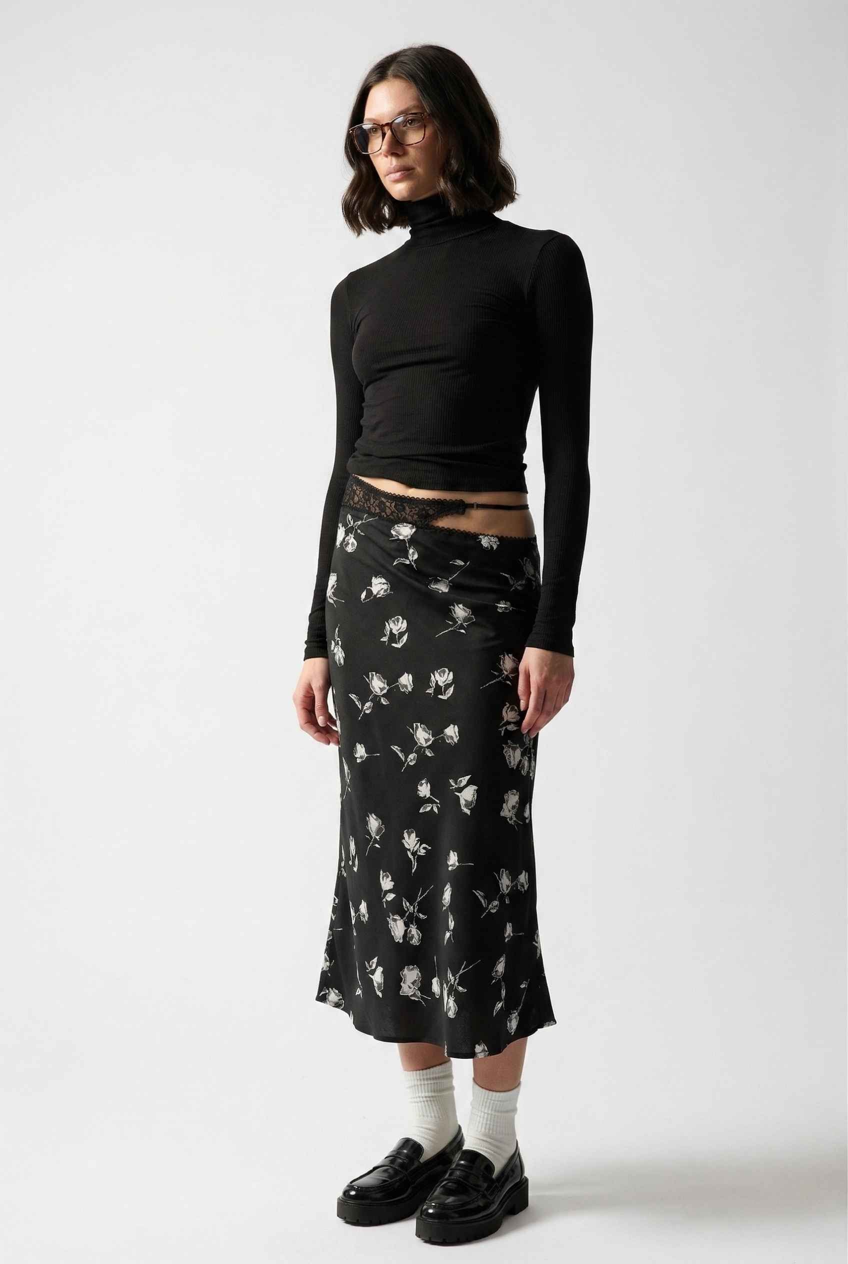Model wearing Raven Skirt in Black Floral Satin Midi Skirt with Lace Waist paired with basic turtle neck and shiny loafers with tall socks