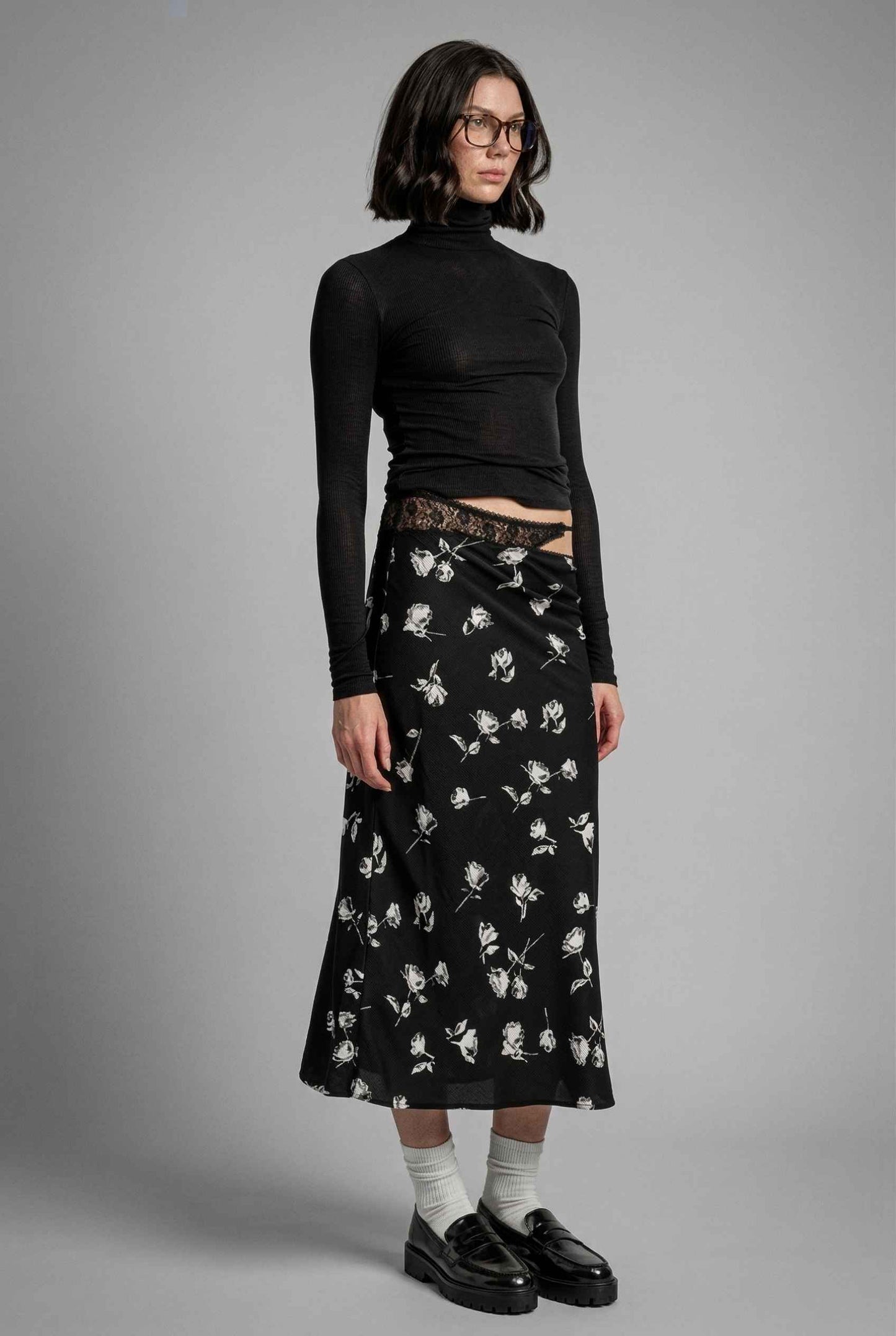 Model wearing Raven Skirt in Black Floral Satin Midi Skirt with Lace Waist paired with basic turtle neck and shiny loafers with tall socks