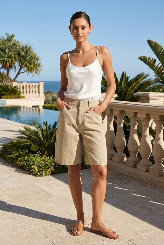 Model wearing Riley Shorts in warm Beige Cotton Denim High-Rise Bermuda paired satin tank top and leather slides luxury vacation pool