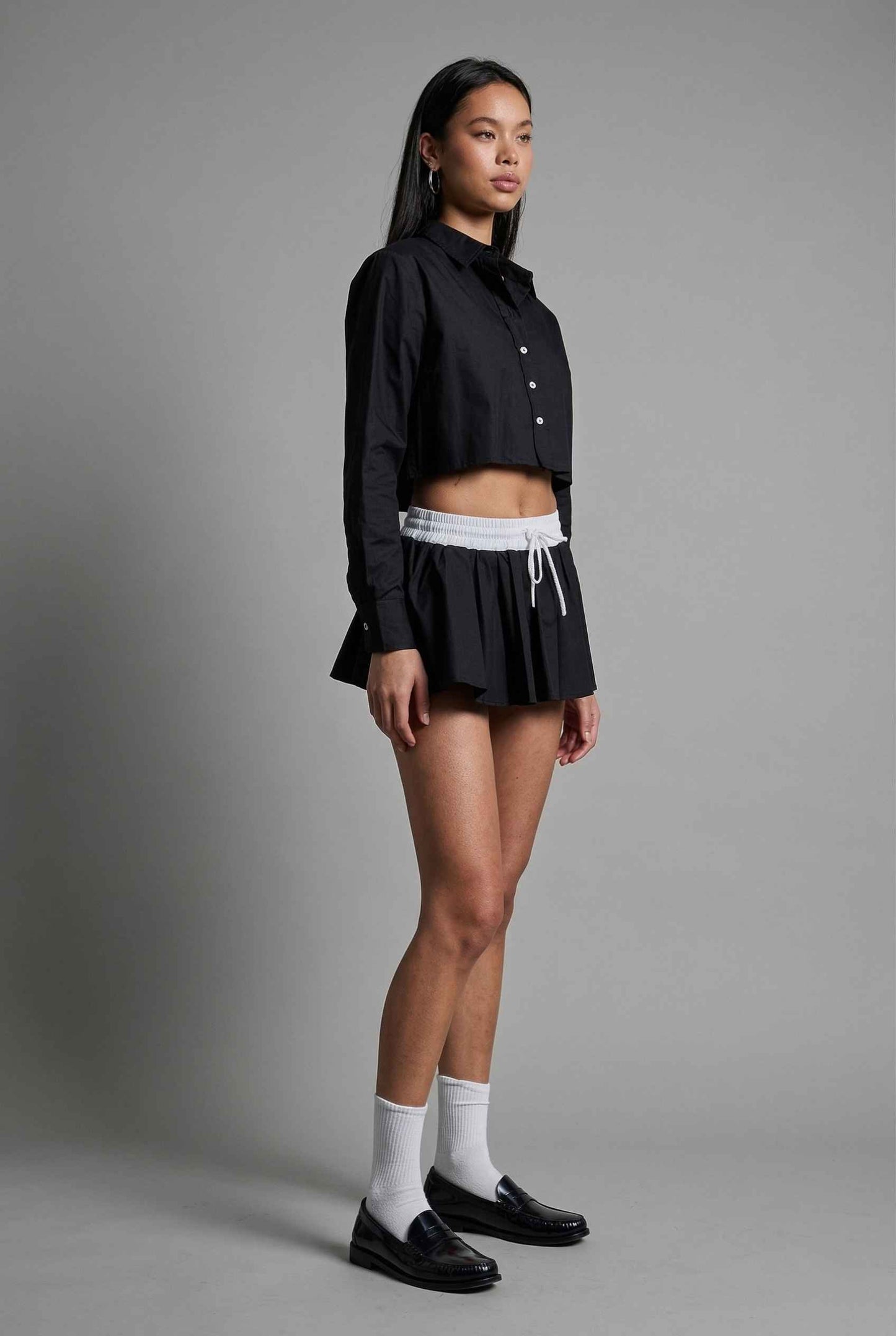 Model wearing Riley Skirt in Black Cotton Poplin Boxer Waist Mini Skirt paired with matching poplin long sleeve button top and shiny loafers with tall socks