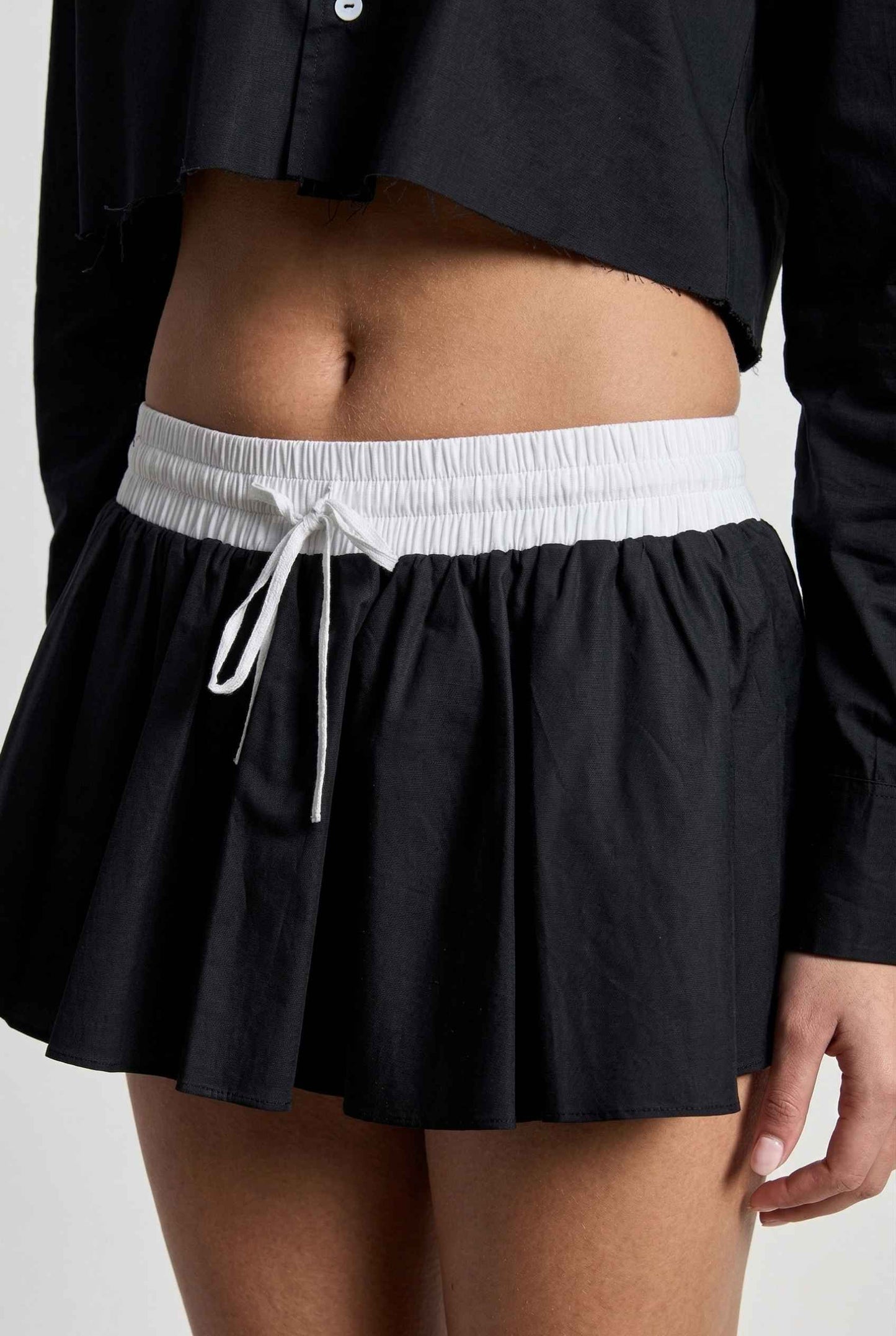Model wearing Riley Skirt in Black Cotton Poplin Boxer Waist Mini Skirt paired with matching poplin long sleeve button top close up tied detail