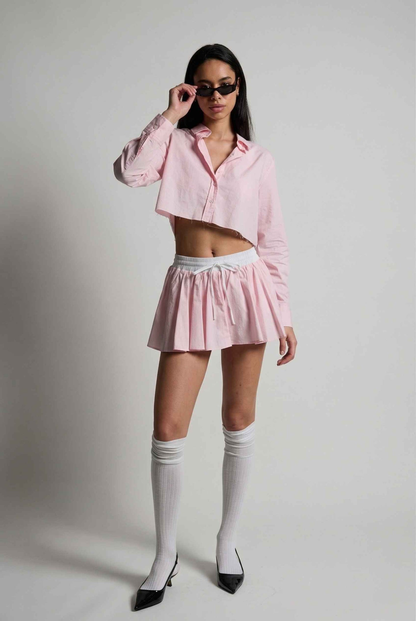 Model wearing Riley Skirt in Pink Cotton Poplin Boxer Waist Mini Skirt paired with matching poplin long sleeve button top and sling back heels with tall socks