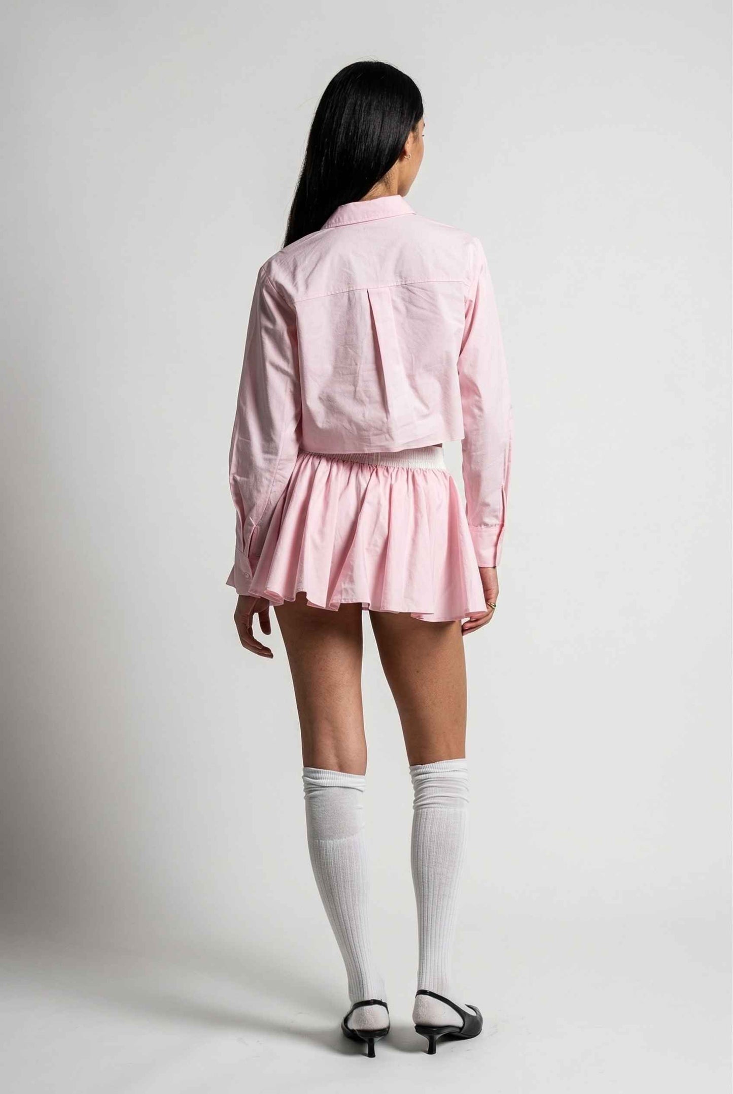 Model wearing Riley Skirt in Pink Cotton Poplin Boxer Waist Mini Skirt paired with matching poplin long sleeve button top and sling back heels with tall socks