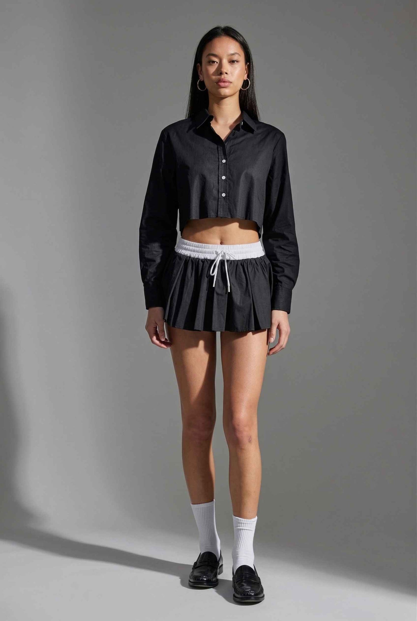 Model wearing Riley Skirt in Black Cotton Poplin Boxer Waist Mini Skirt paired with matching poplin long sleeve button top and shiny loafers with tall socks