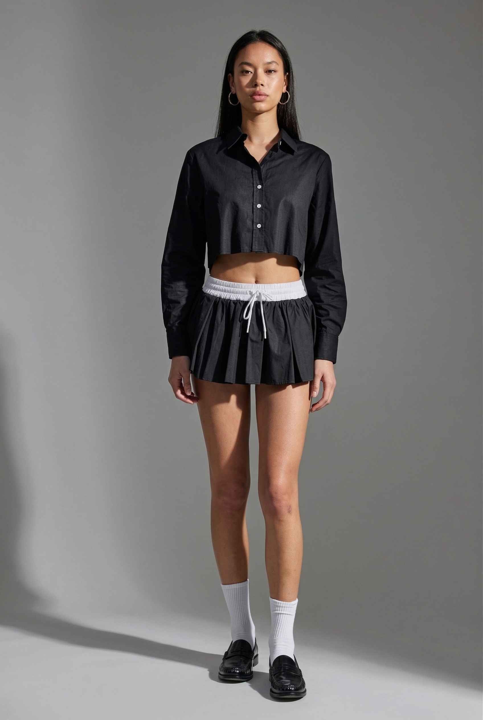 Model wearing Riley Skirt in Black Cotton Poplin Boxer Waist Mini Skirt paired with matching poplin long sleeve button top and shiny loafers with tall socks