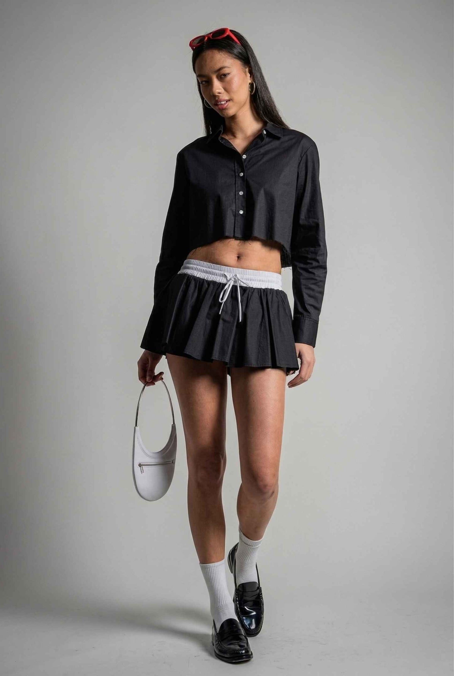 Model wearing Riley Skirt in Black Cotton Poplin Boxer Waist Mini Skirt paired with matching poplin long sleeve button top and shiny loafers with tall socks