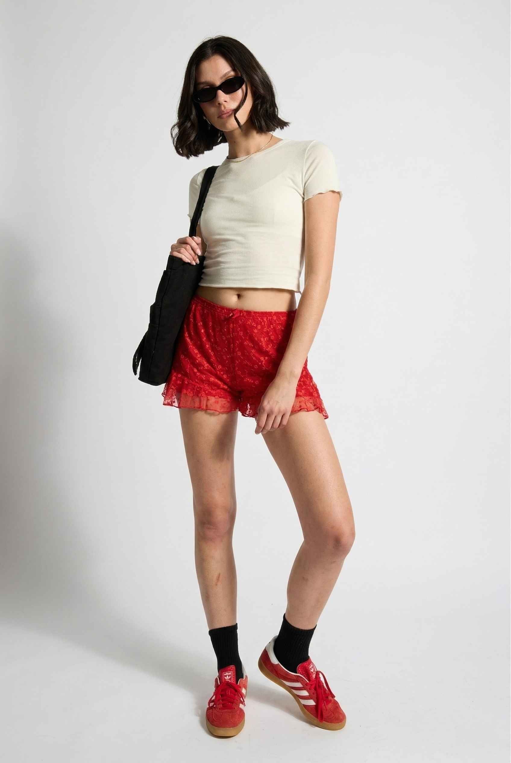 Model wearing Ruby Shorts in Red Lace Ruffle Bloomer Shorts with Bow paired with cropped t-shirt and casual sneakers with socks