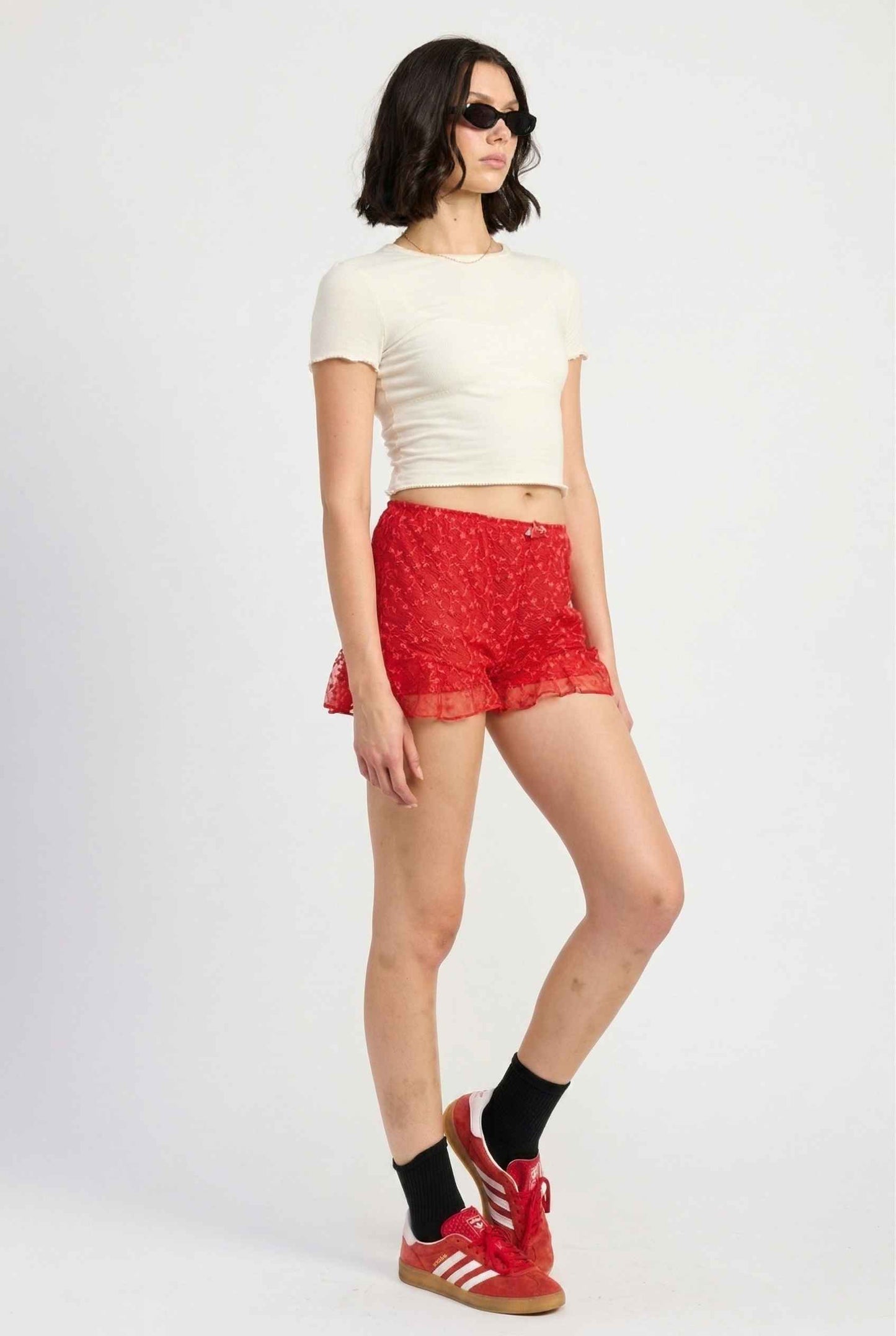 Model wearing Ruby Shorts in Red Lace Ruffle Bloomer Shorts with Bow paired with cropped t-shirt and casual sneakers with socks