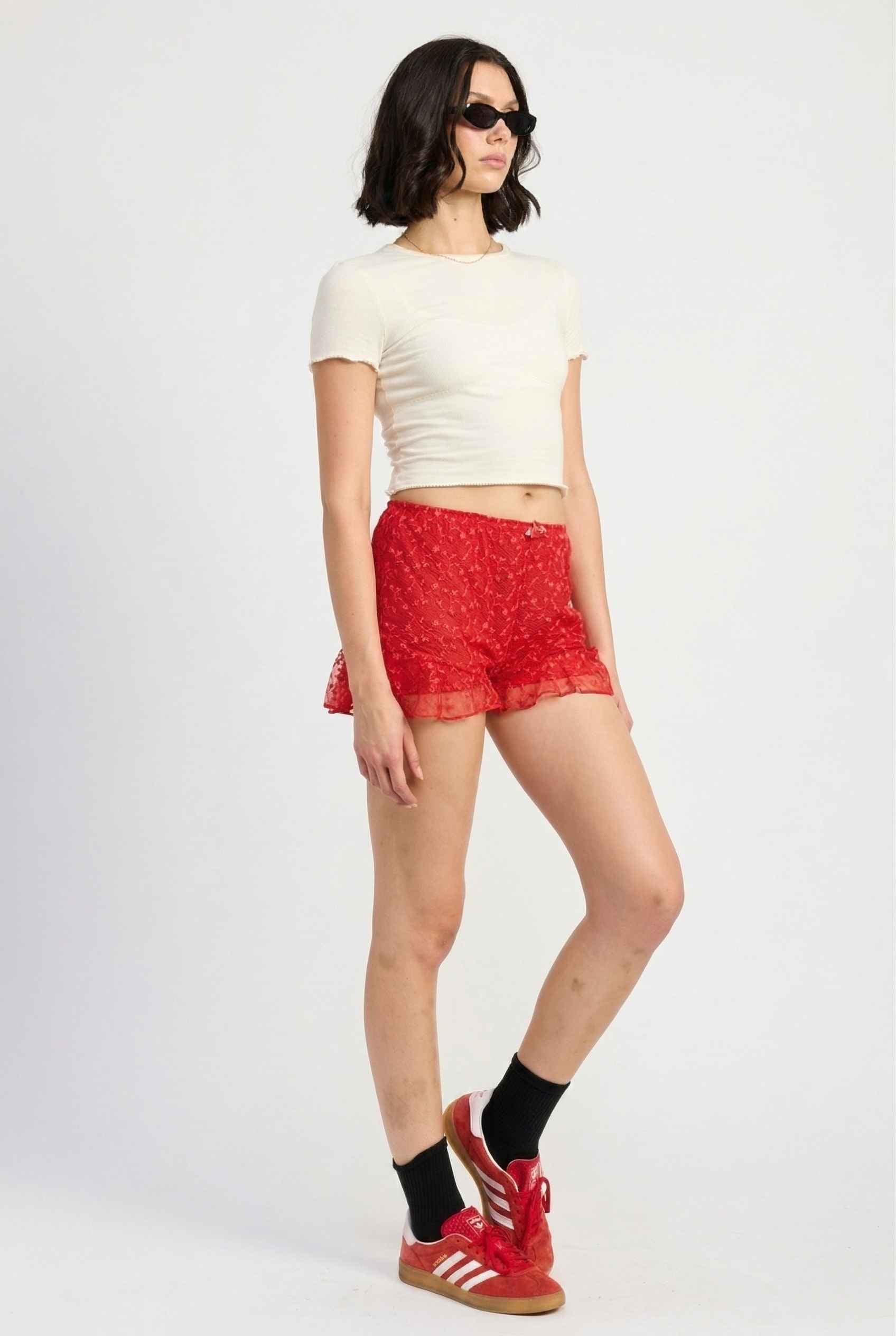 Model wearing Ruby Shorts in Red Lace Ruffle Bloomer Shorts with Bow paired with cropped t-shirt and casual sneakers with socks