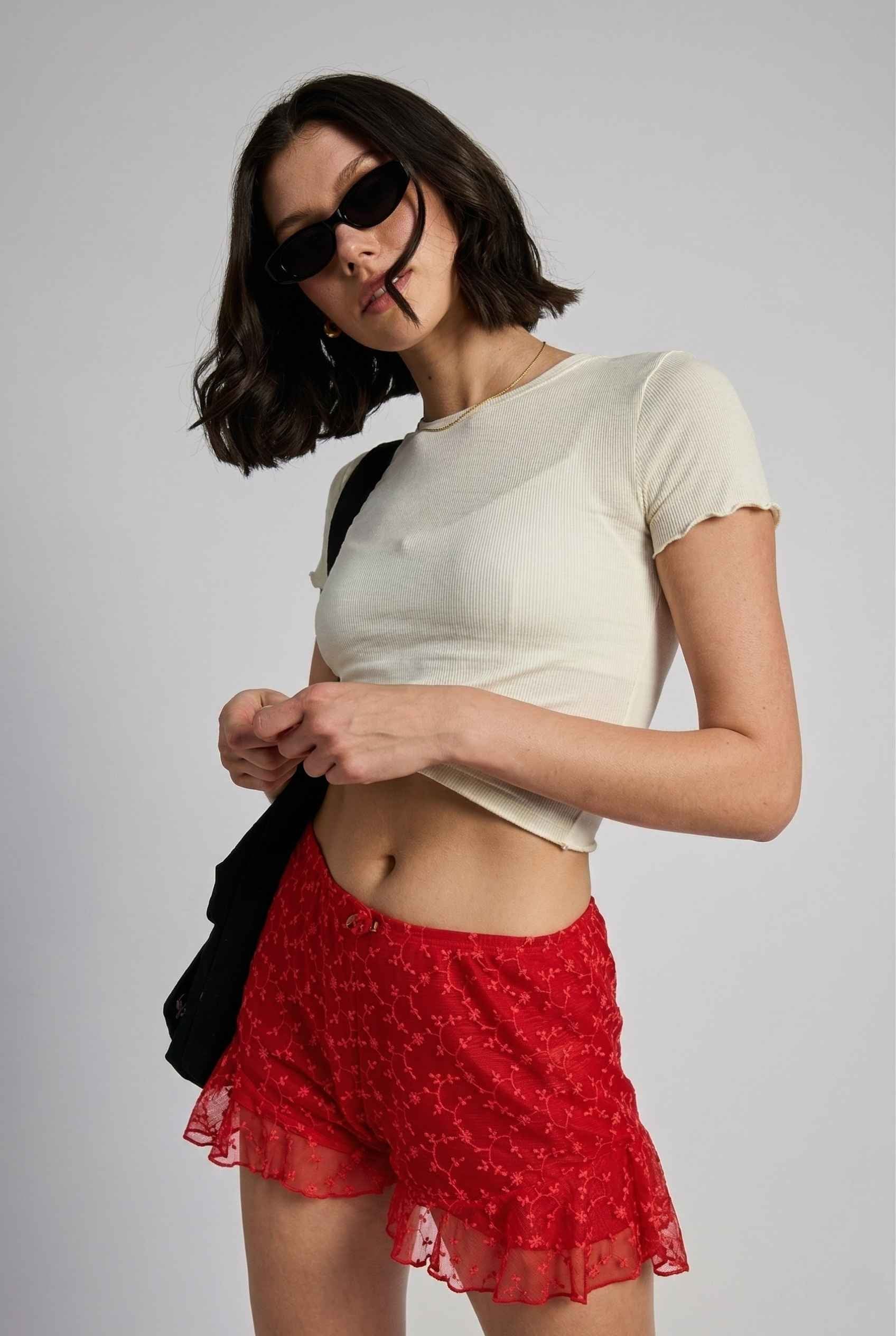 Model wearing Ruby Shorts in Red Lace Ruffle Bloomer Shorts with Bow paired with cropped t-shirt and shoulder bag