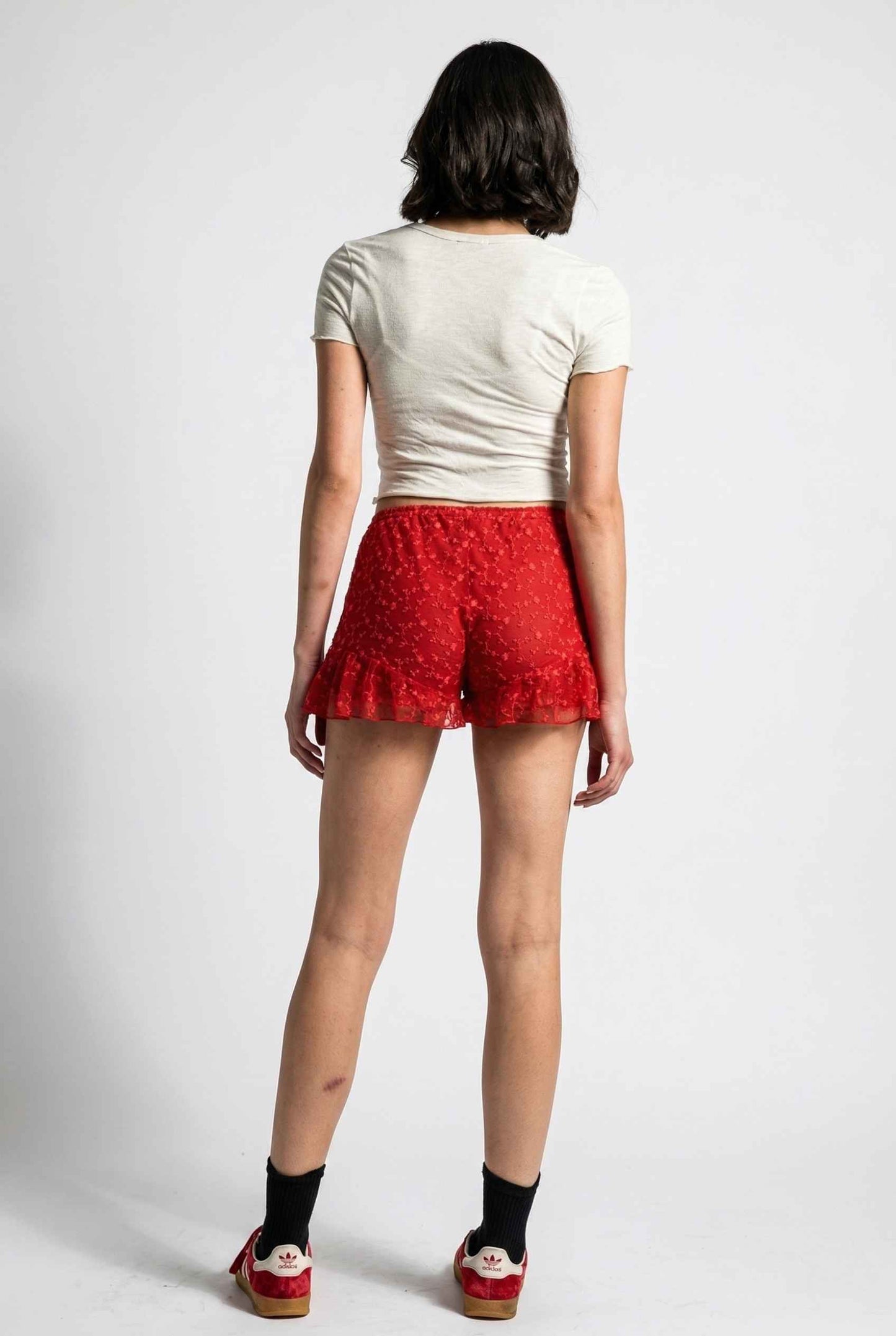 Model wearing Ruby Shorts in Red Lace Ruffle Bloomer Shorts with Bow paired with cropped t-shirt and casual sneakers with socks
