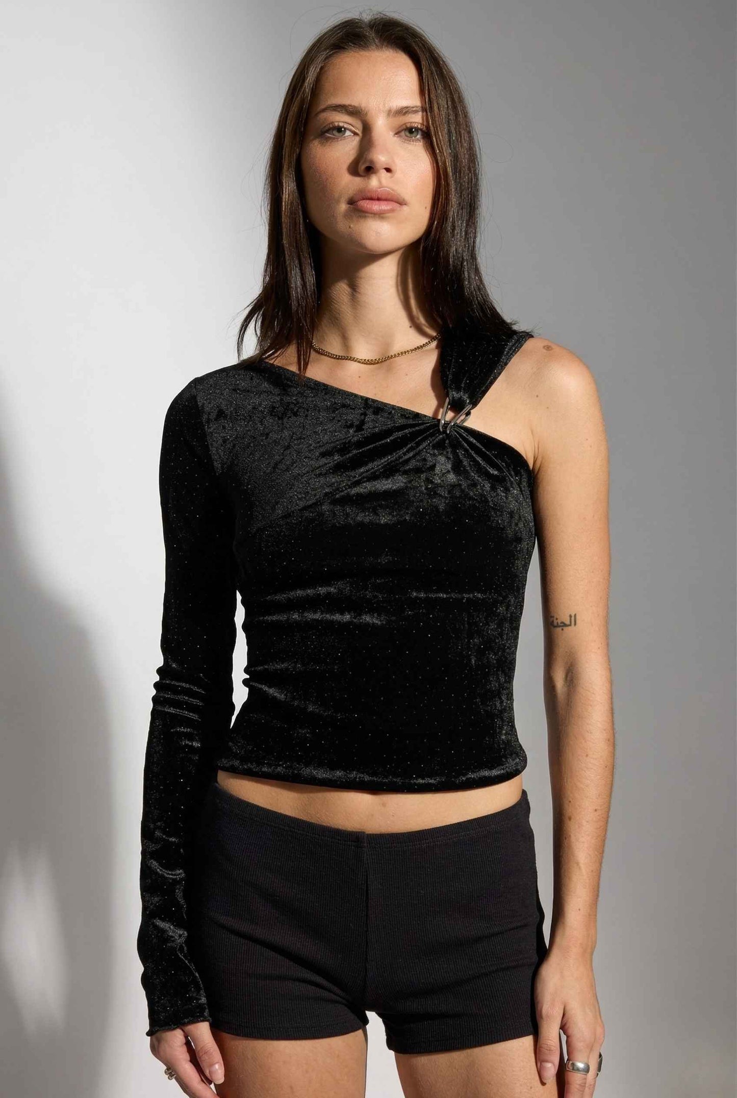 Woman wearing a Selene Top Black Shimmer Velvet Asymmetrical Long Sleeve and black shorts against a gray background