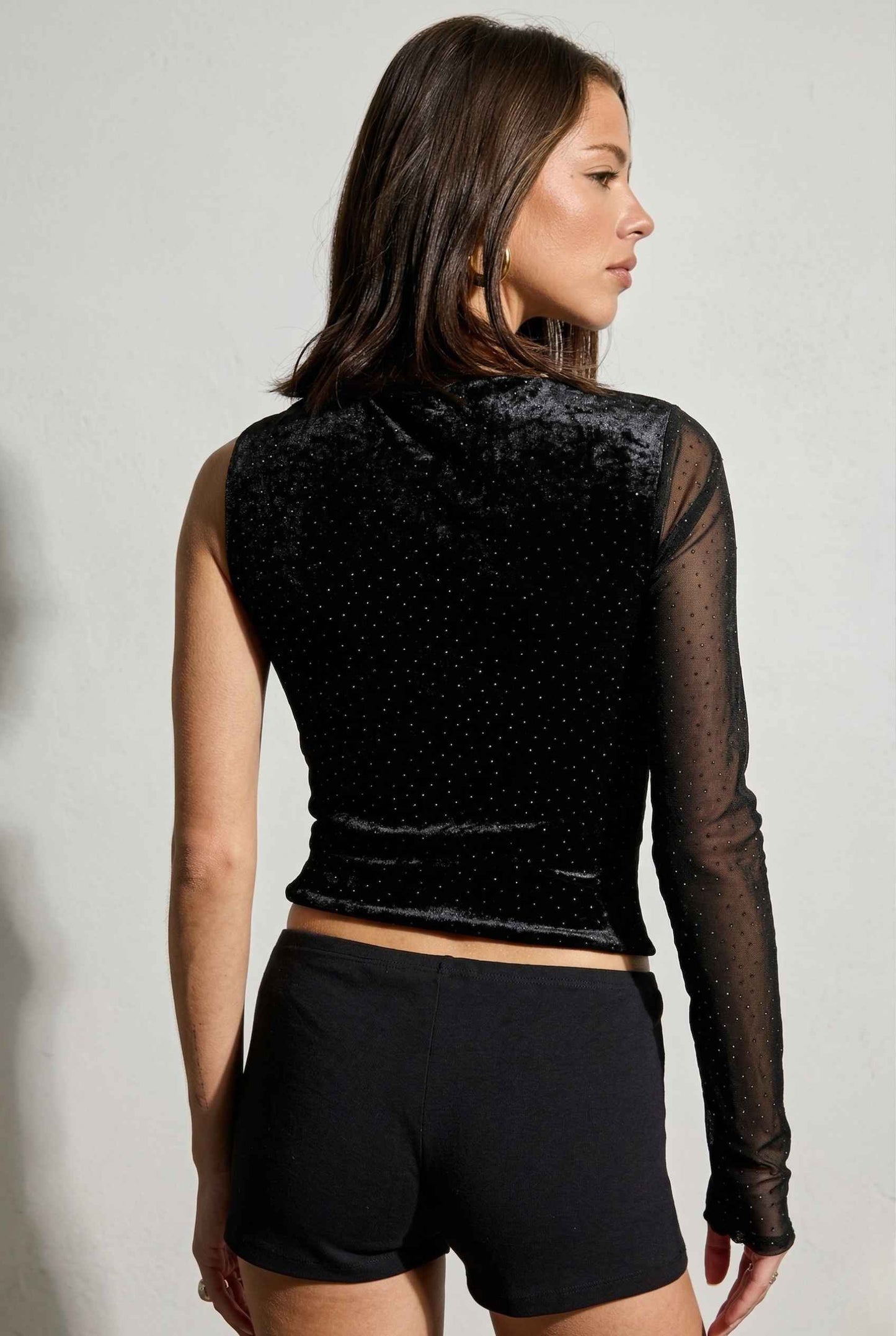 Woman wearing a Selene Top Black Shimmer Velvet Asymmetrical Long Sleeve with sheer panels and black shorts against a plain background