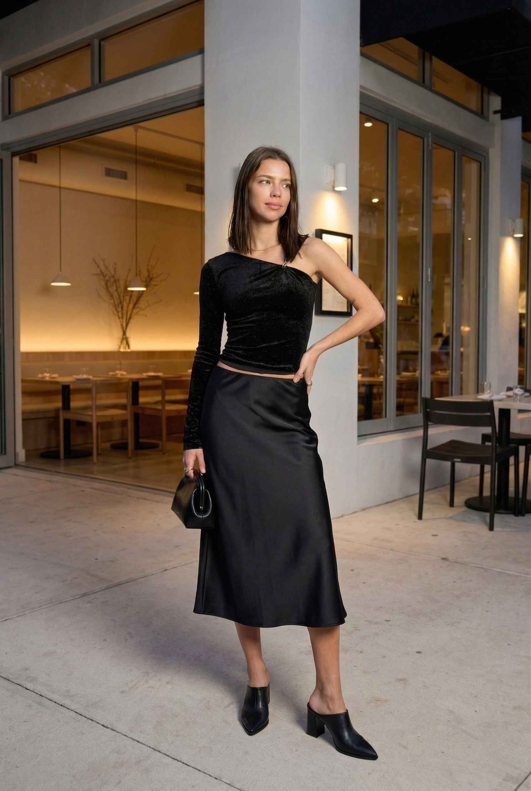 Woman in a Selene Top Black Shimmer Velvet Asymmetrical Long Sleeve standing outside a building with a modern interior.