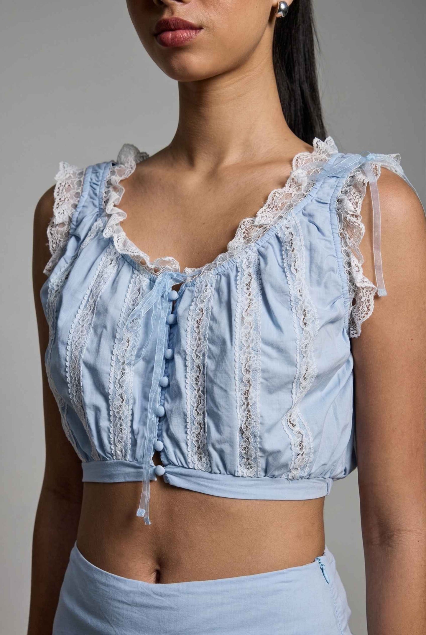 Close up of a woman wearing a Seraphina Top Light Blue & White Cotton Lace Ruffle Bralette on a plain background. 