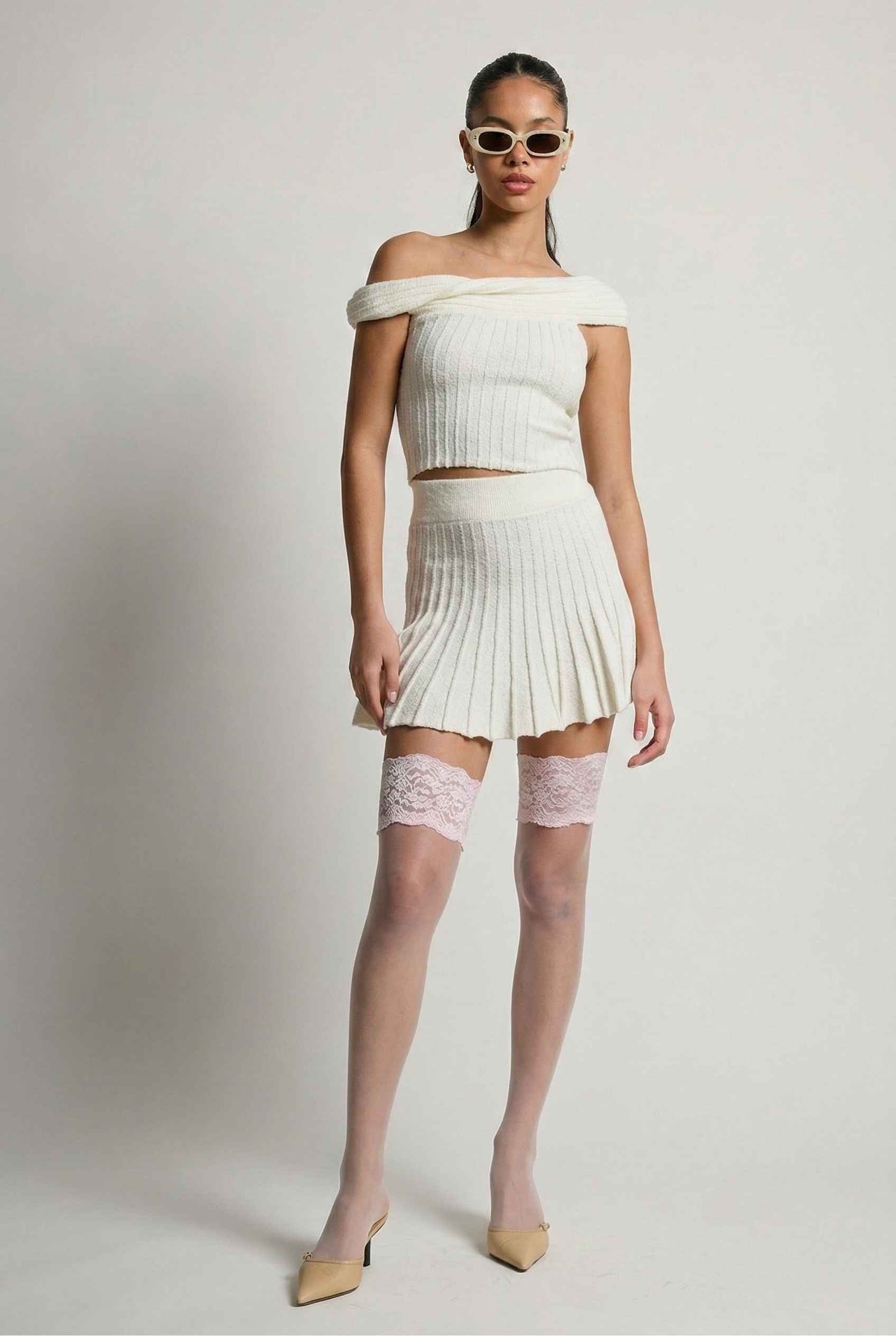 Model wearing Seraphina Top in White Ribbed Knit Twist-Front Off-Shoulder Top with matching mini skirt, lace tights and heels on plain background