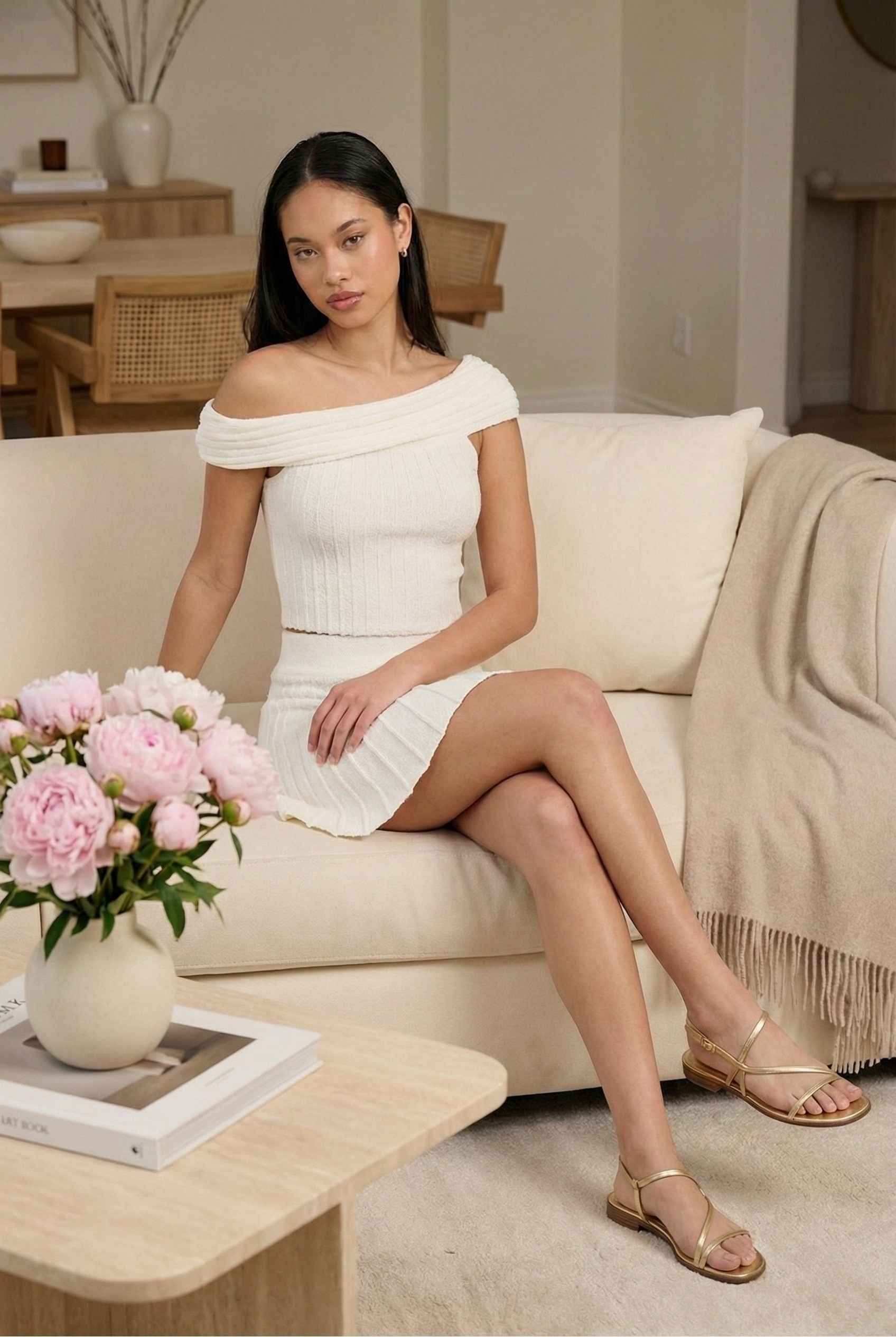 Model wearing Seraphina Top in White Ribbed Knit Twist-Front Off-Shoulder Top with matching mini skirt and sandals posing in minimalist luxury living room