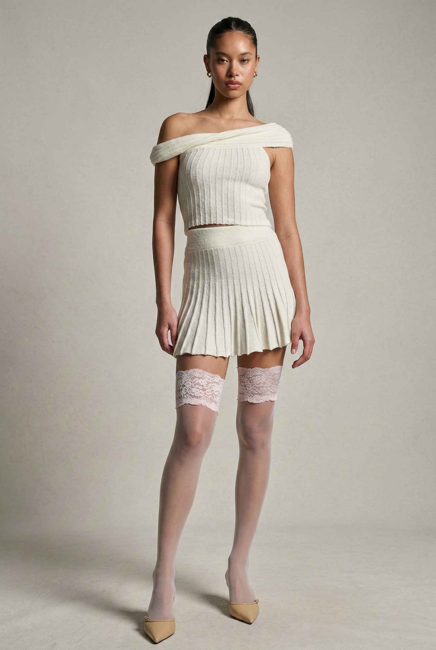 Model wearing Seraphina Top in White Ribbed Knit Twist-Front Off-Shoulder Top with matching mini skirt, lace tights and heels on plain background