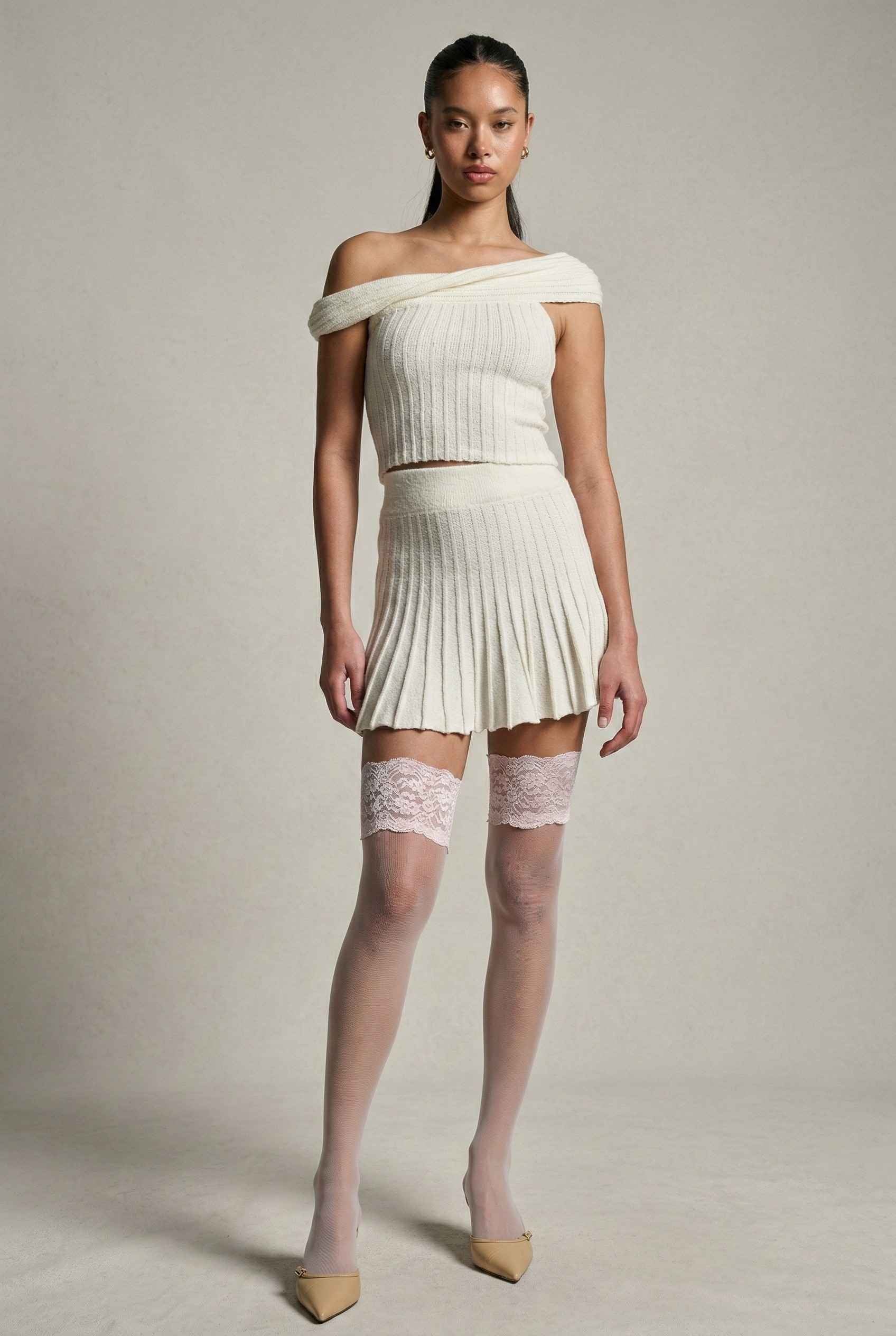 Model wearing Seraphina Top in White Ribbed Knit Twist-Front Off-Shoulder Top with matching mini skirt, lace tights and heels on plain background