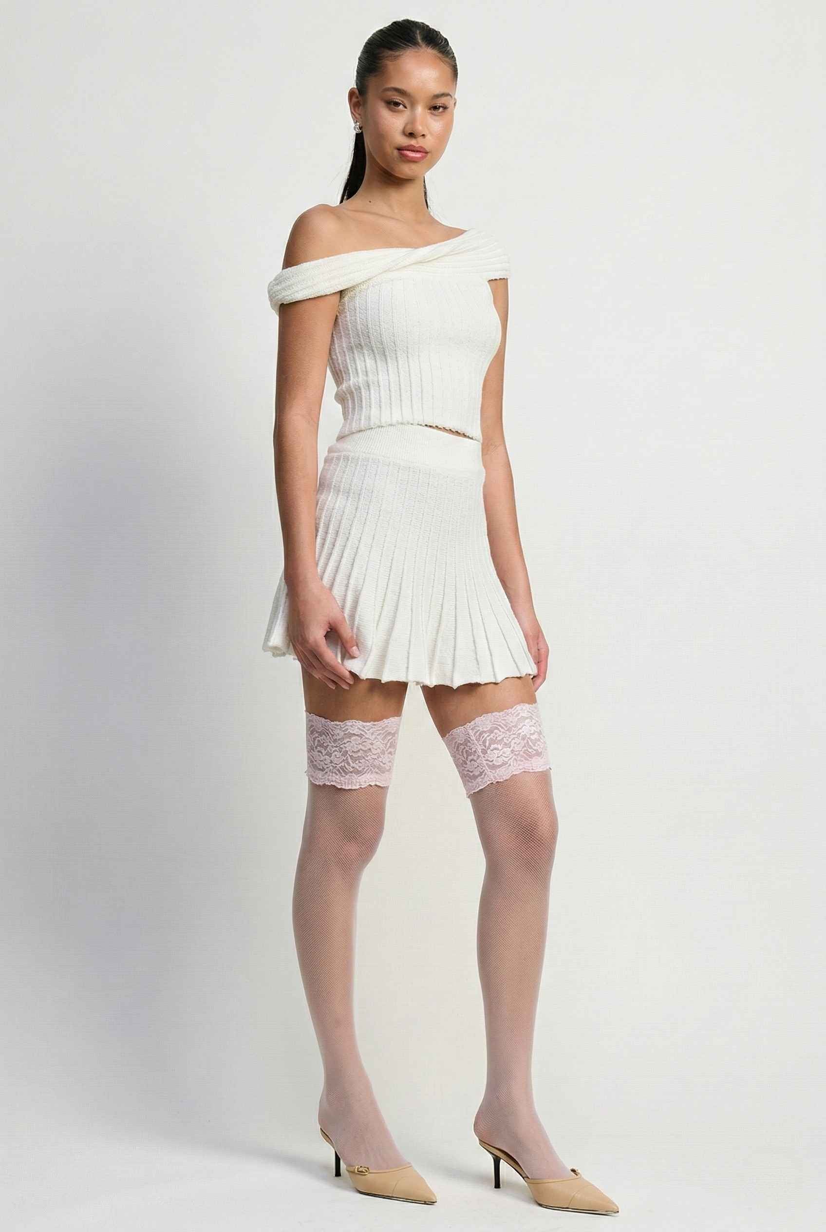 Model wearing Seraphina Top in White Ribbed Knit Twist-Front Off-Shoulder Top with matching mini skirt, lace tights and heels on plain background