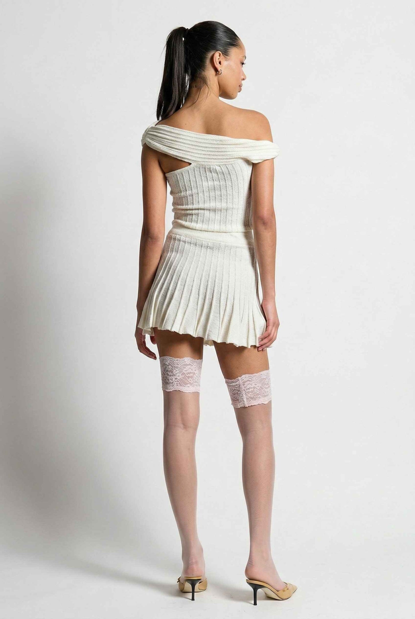 Model wearing Seraphina Top in White Ribbed Knit Twist-Front Off-Shoulder Top with matching mini skirt, lace tights and heels on plain background