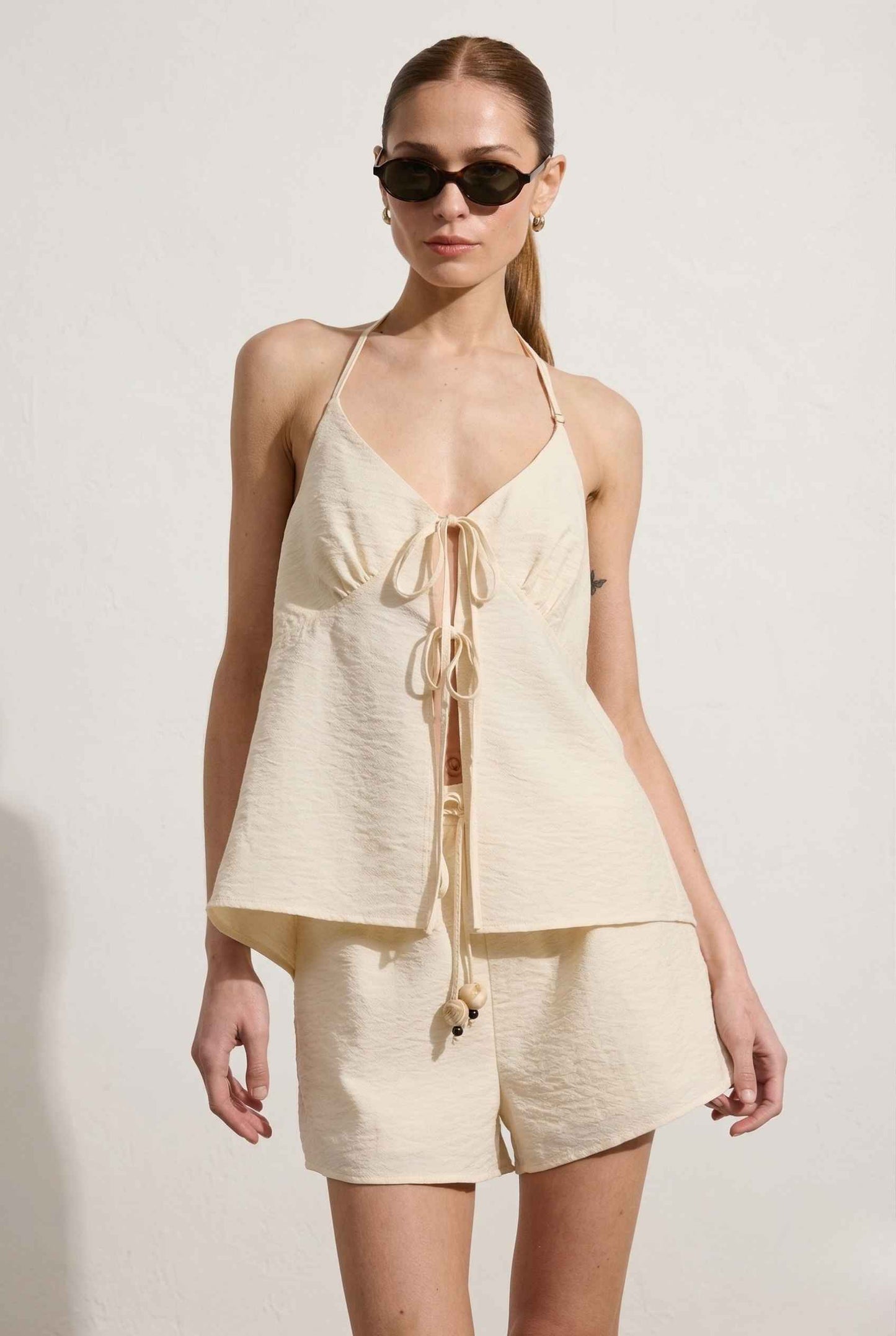 Woman wearing a Sienna Top Cream Tie-Front V-Neck Halter Tank and shorts set with sunglasses on a plain background