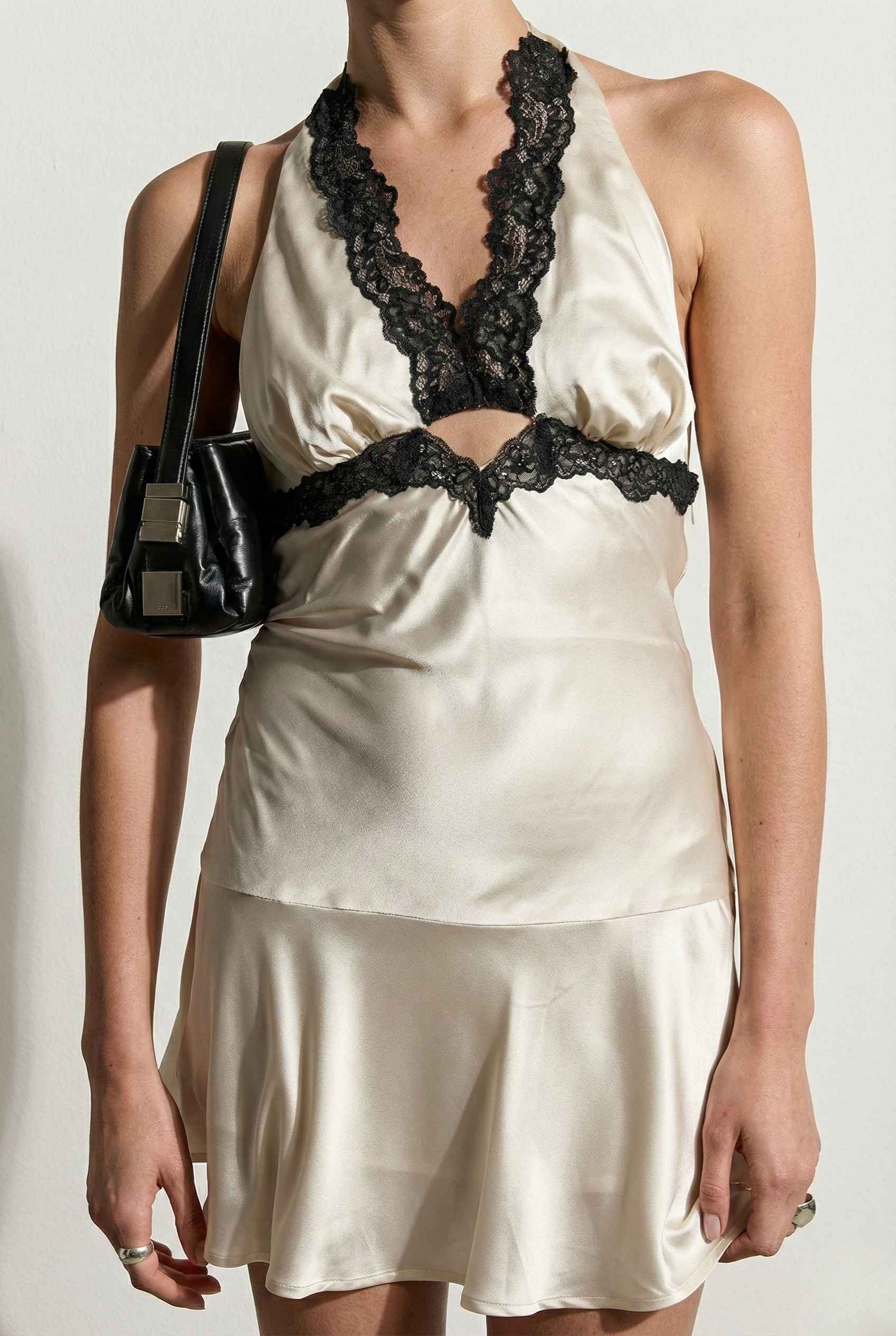 Woman wearing a Simone Mini Ivory Satin Halter Lace Trim Dress with black lace details and holding a black handbag.
