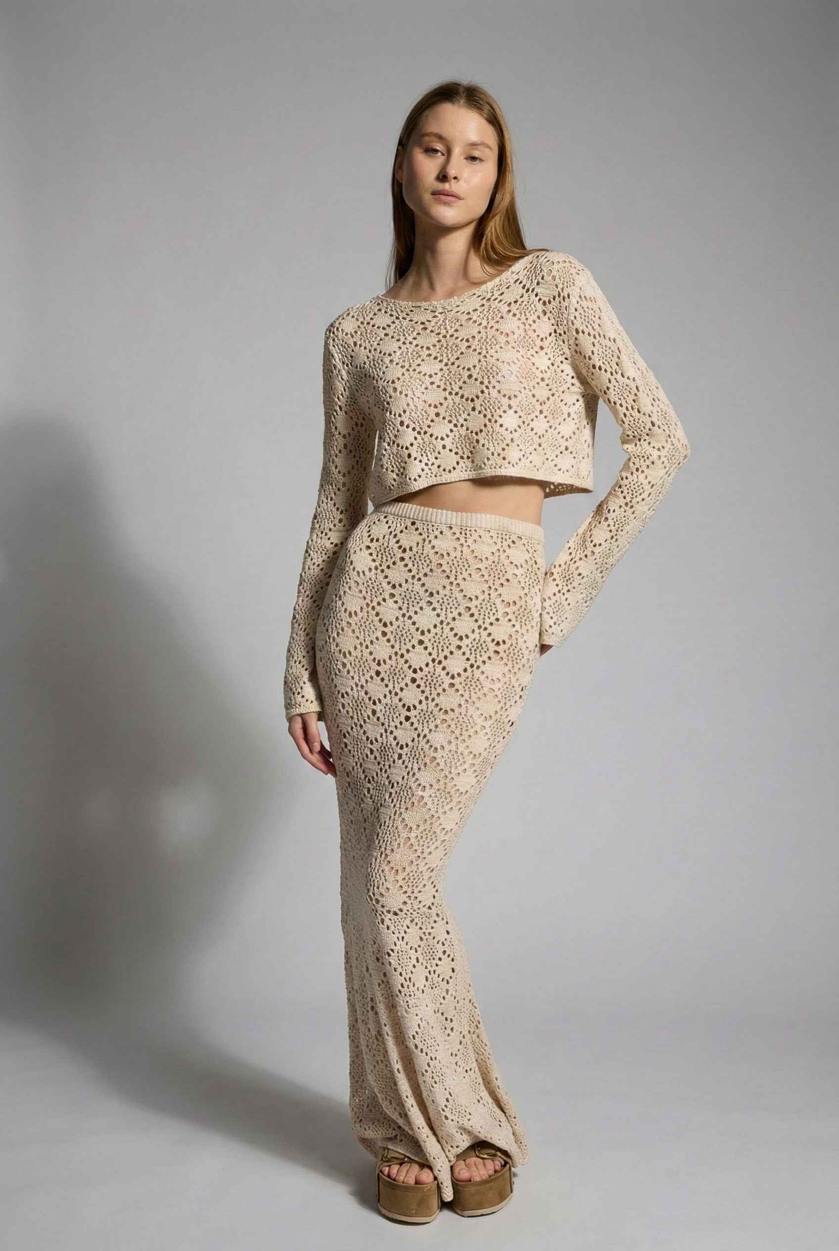 Model wearing Siren Skirt Open-Knit Crochet Mermaid Maxi Skirt in Sand Cream with matching crop top and platform sandals on plain background