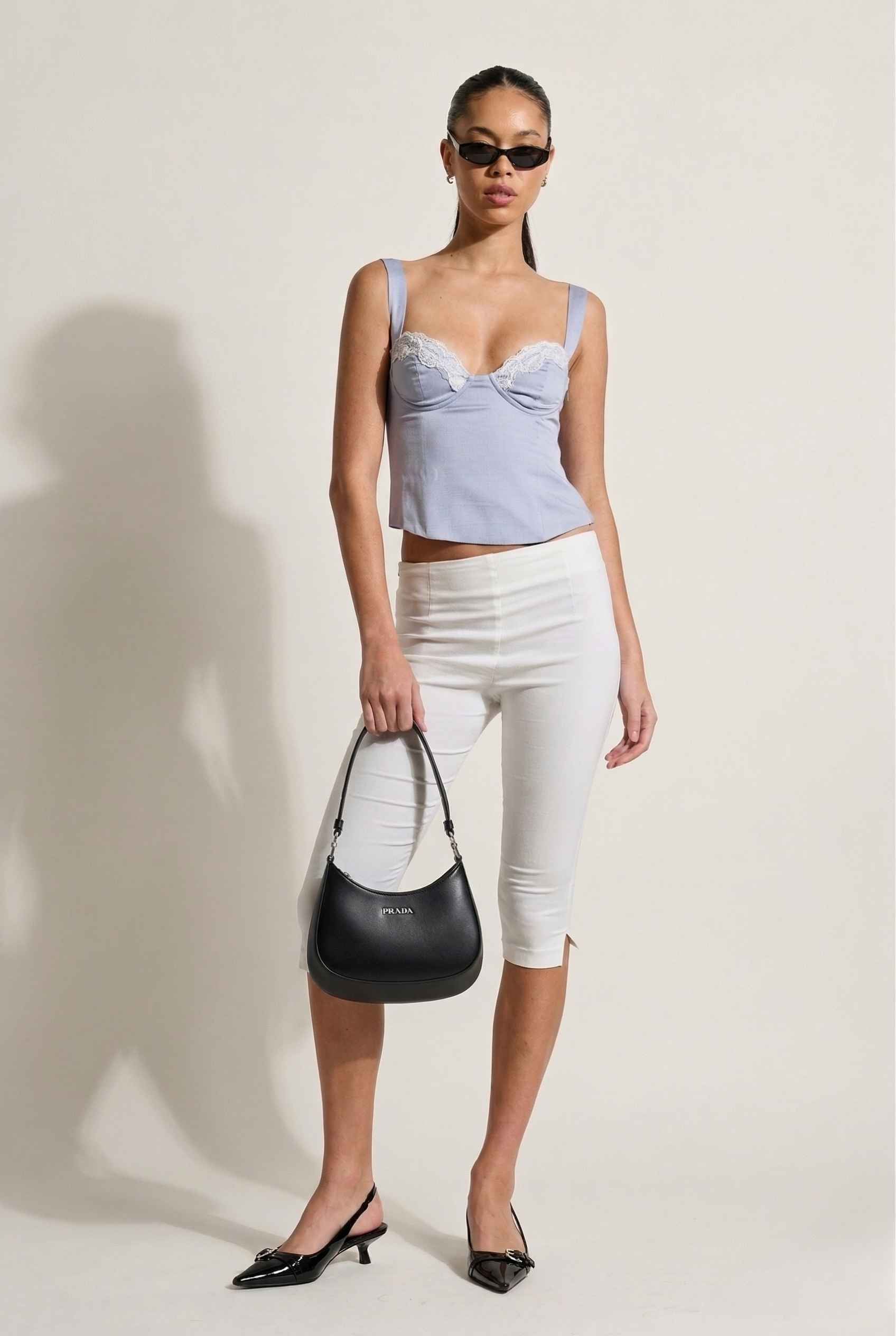 Woman wearing a Sophie Top Blue Pastel Satin Corset Cami with White Lace Trim and white pants, holding a black handbag against a plain background
