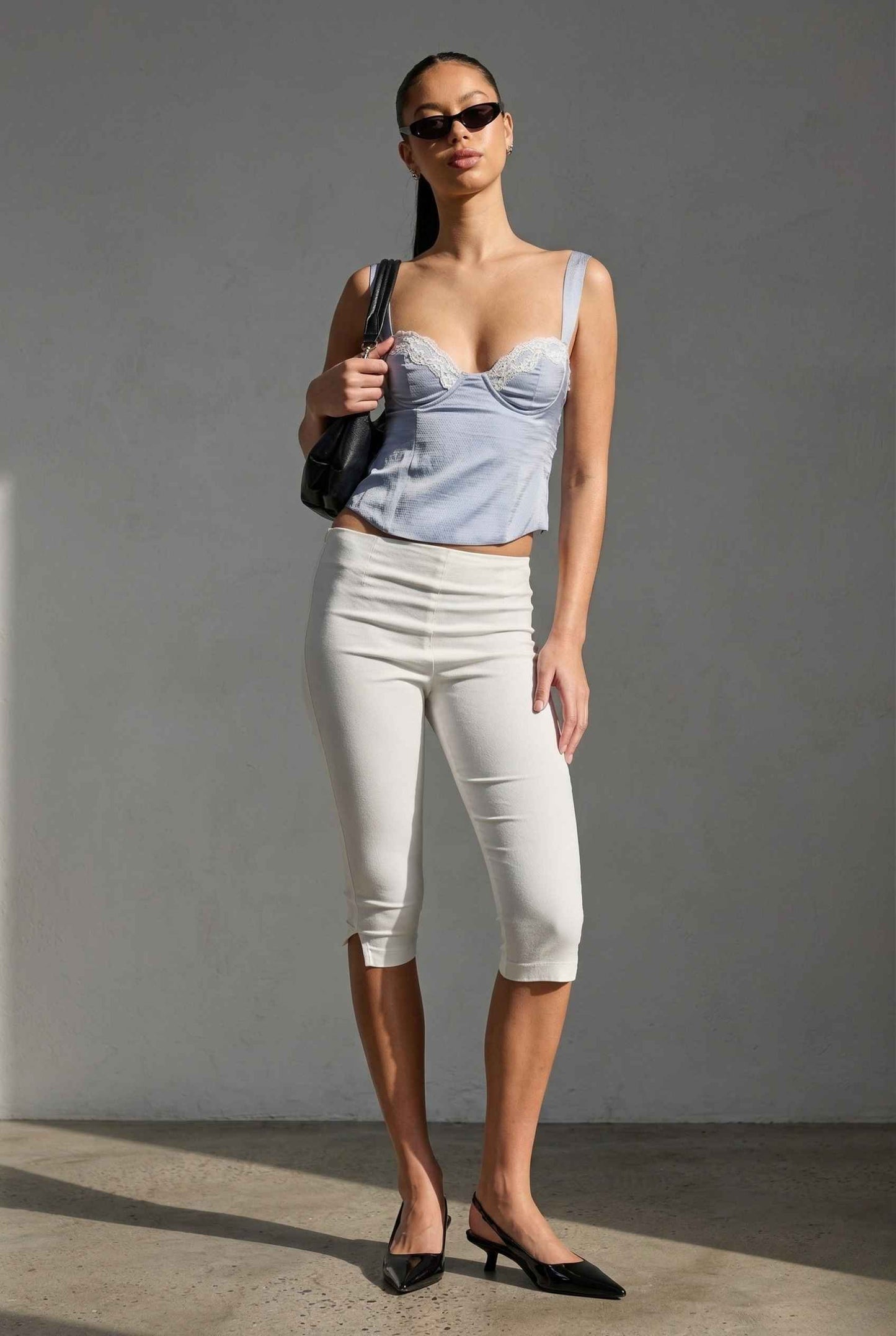 Woman wearing a Sophie Top Blue Pastel Satin Corset Cami with White Lace Trim and white pants against a gray background