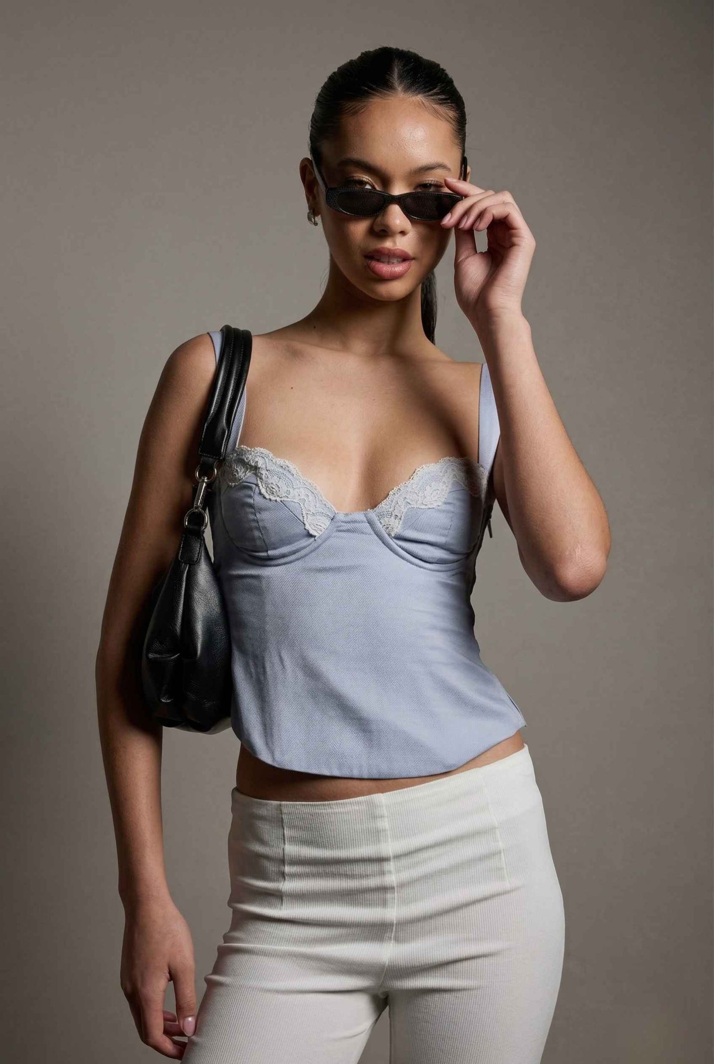 Woman wearing a Sophie Top Blue Pastel Satin Corset Cami with White Lace Trim and white pants, holding sunglasses and a black handbag against a gray background