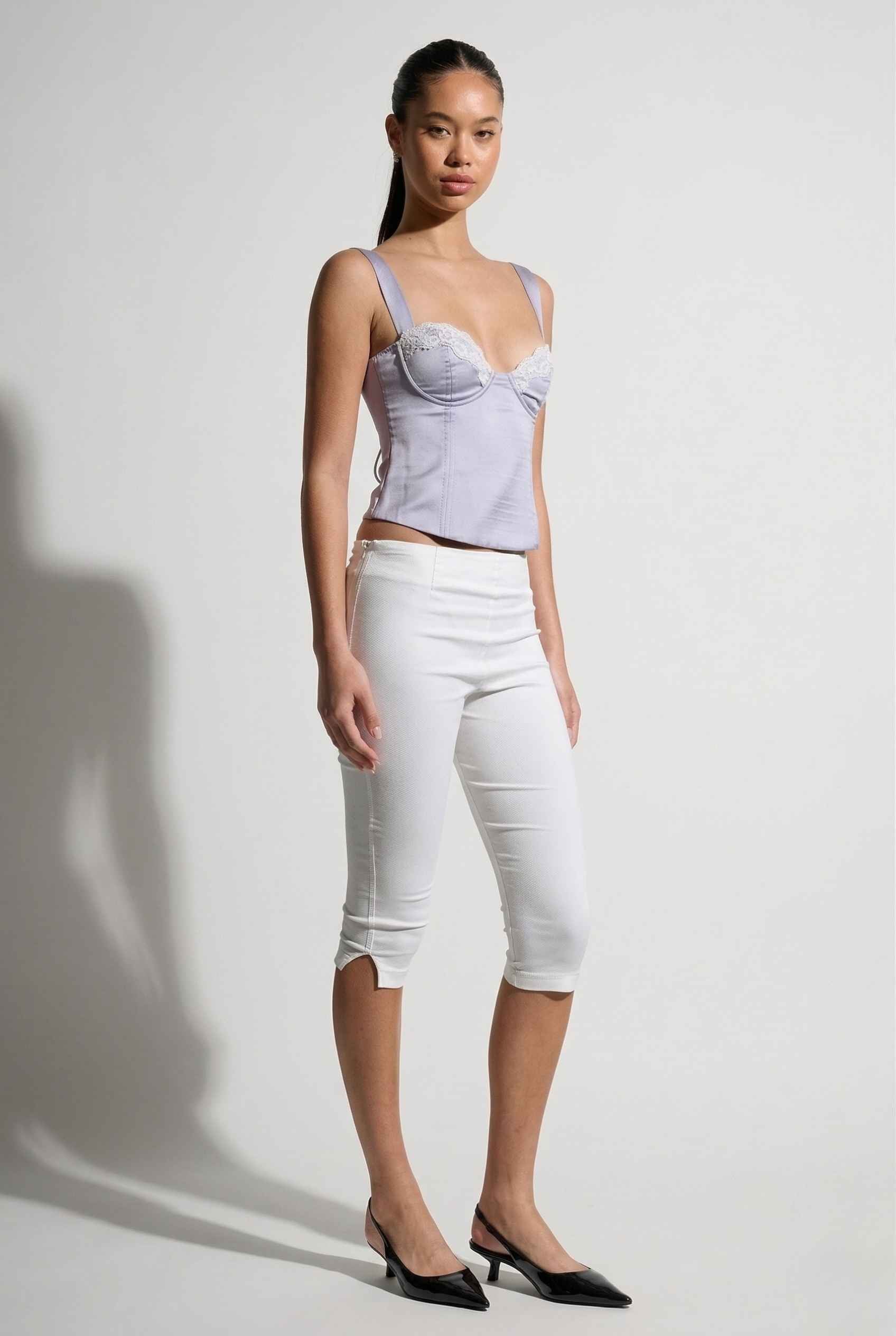 Woman wearing a Sophie Top Blue Pastel Satin Corset Cami with White Lace Trim and white pants against a plain background