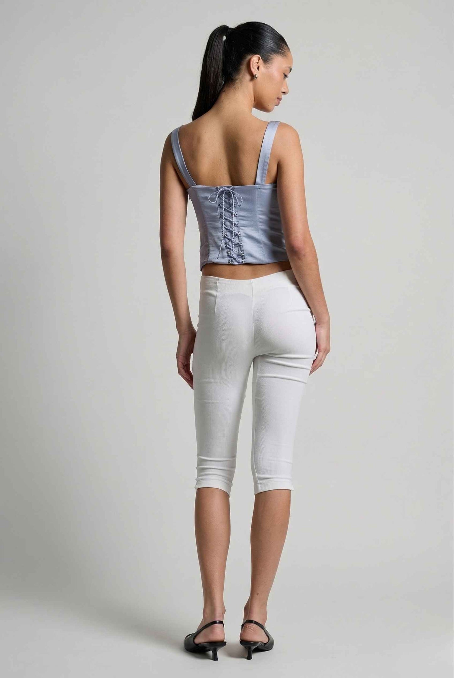 Woman wearing a Sophie Top Blue Pastel Satin Corset Cami with White Lace Trim and white capri pants on a plain background