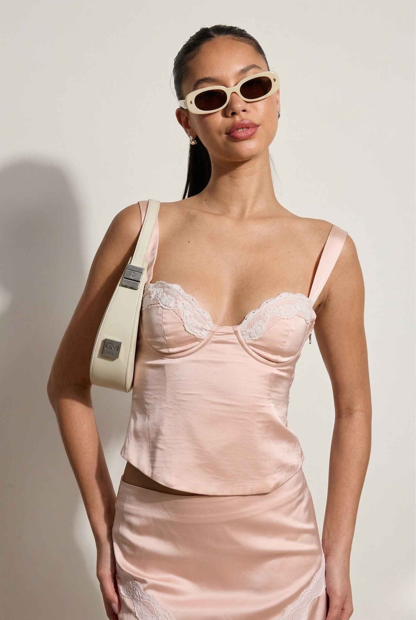 Woman wearing a Sophie Top Pink Pastel Satin Corset Cami with White Lace Trim with lace details, holding a beige handbag against a plain background.