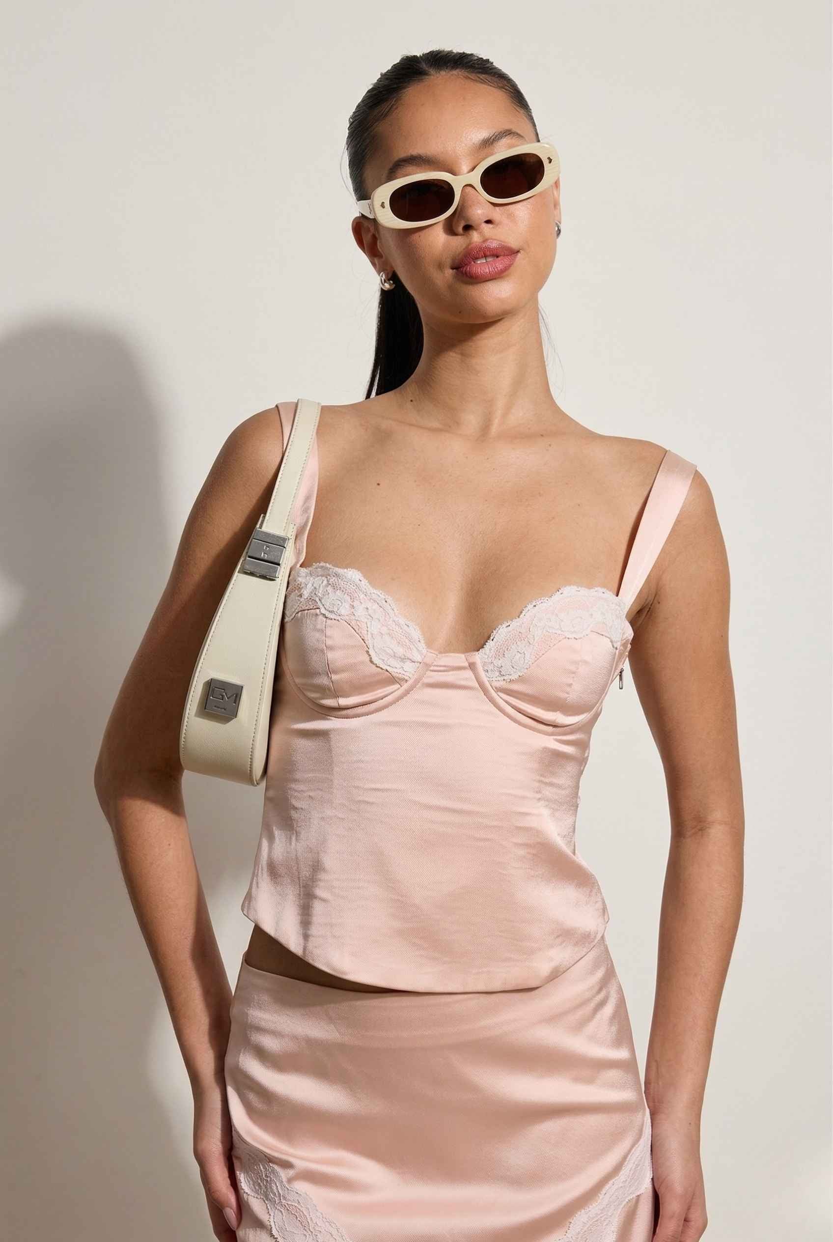 Woman wearing a Sophie Top Pink Pastel Satin Corset Cami with White Lace Trim with lace details, holding a beige handbag against a plain background.