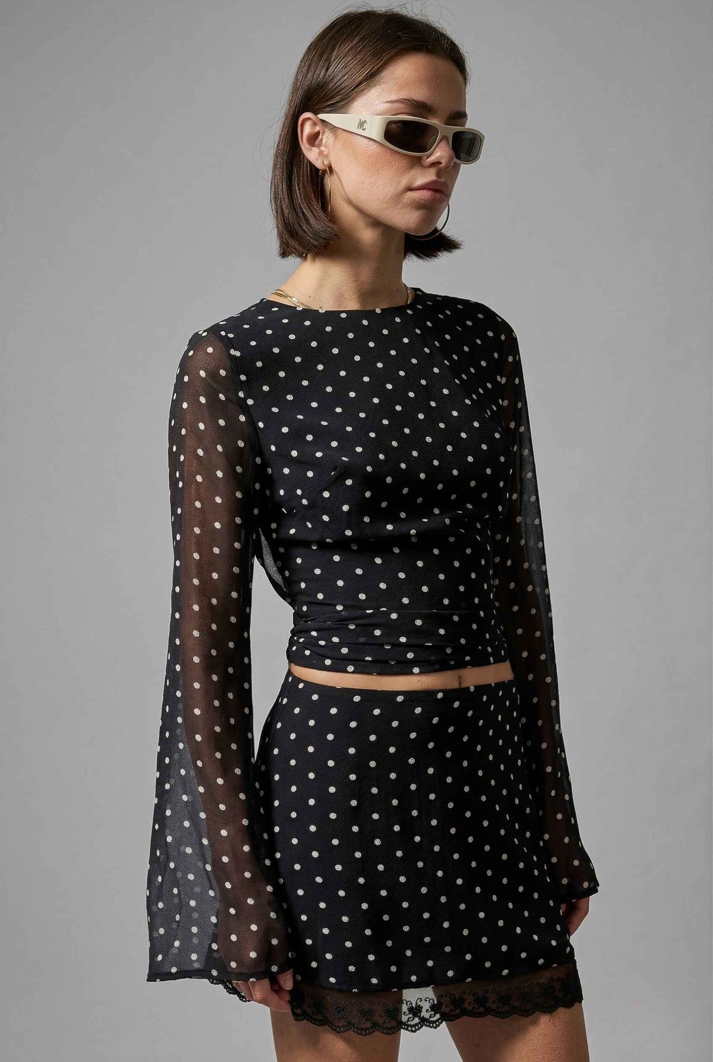 Woman wearing a Stella Top Black & White Polka Dot Sheer Long Sleeve with sheer long sleeves on a gray background
