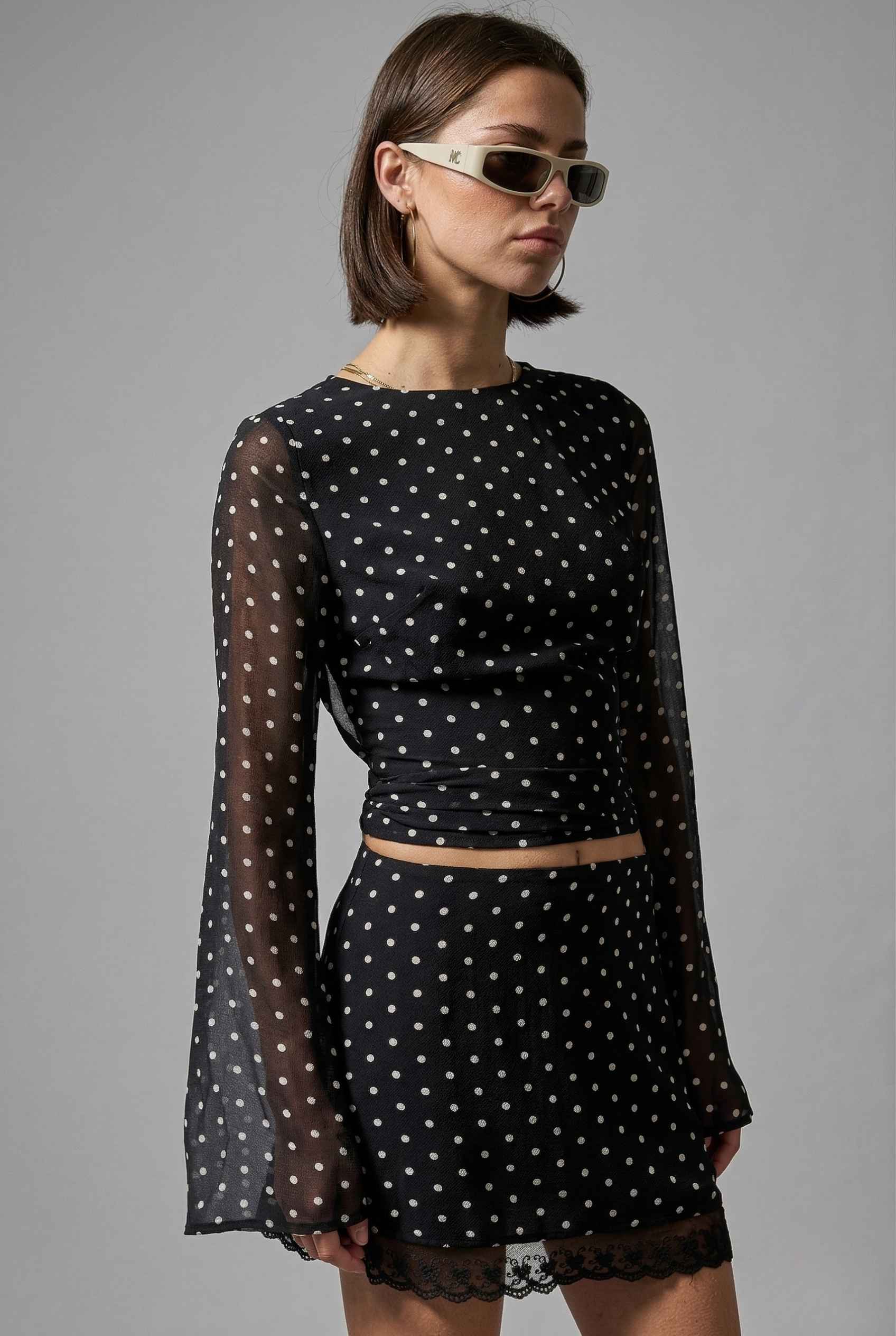 Woman wearing a Stella Top Black & White Polka Dot Sheer Long Sleeve with sheer long sleeves on a gray background