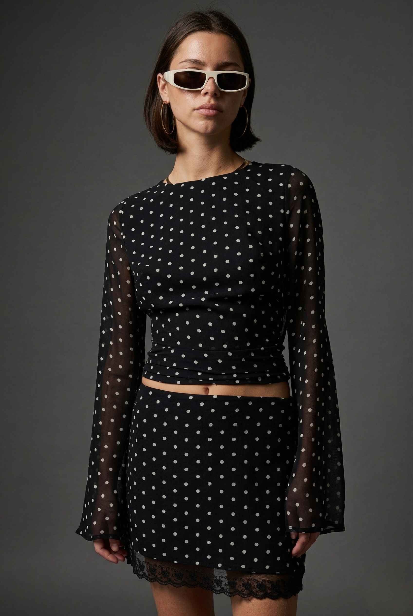 Woman wearing a Stella Top Black & White Polka Dot Sheer Long Sleeve with sheer sleeves against a dark background