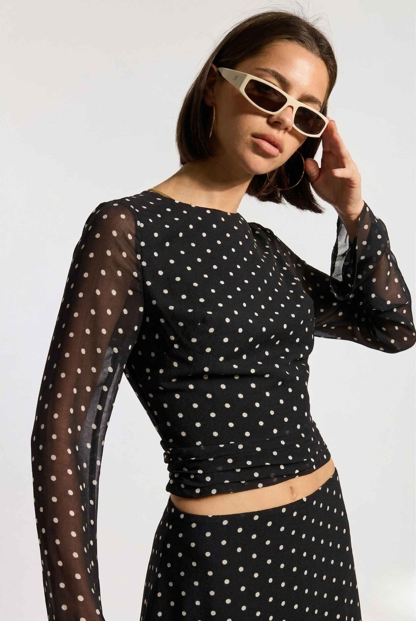 Woman wearing a Stella Top Black & White Polka Dot Sheer Long Sleeve with sunglasses on a white background