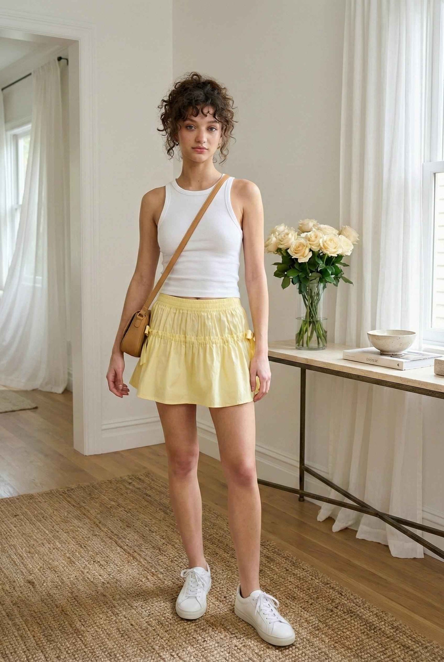 Sunnie Skirt in Soft Yellow Poplin Mini Skirt with Bows paired with ribbed knit tank and sneakers with crossbody purse posing in luxury home