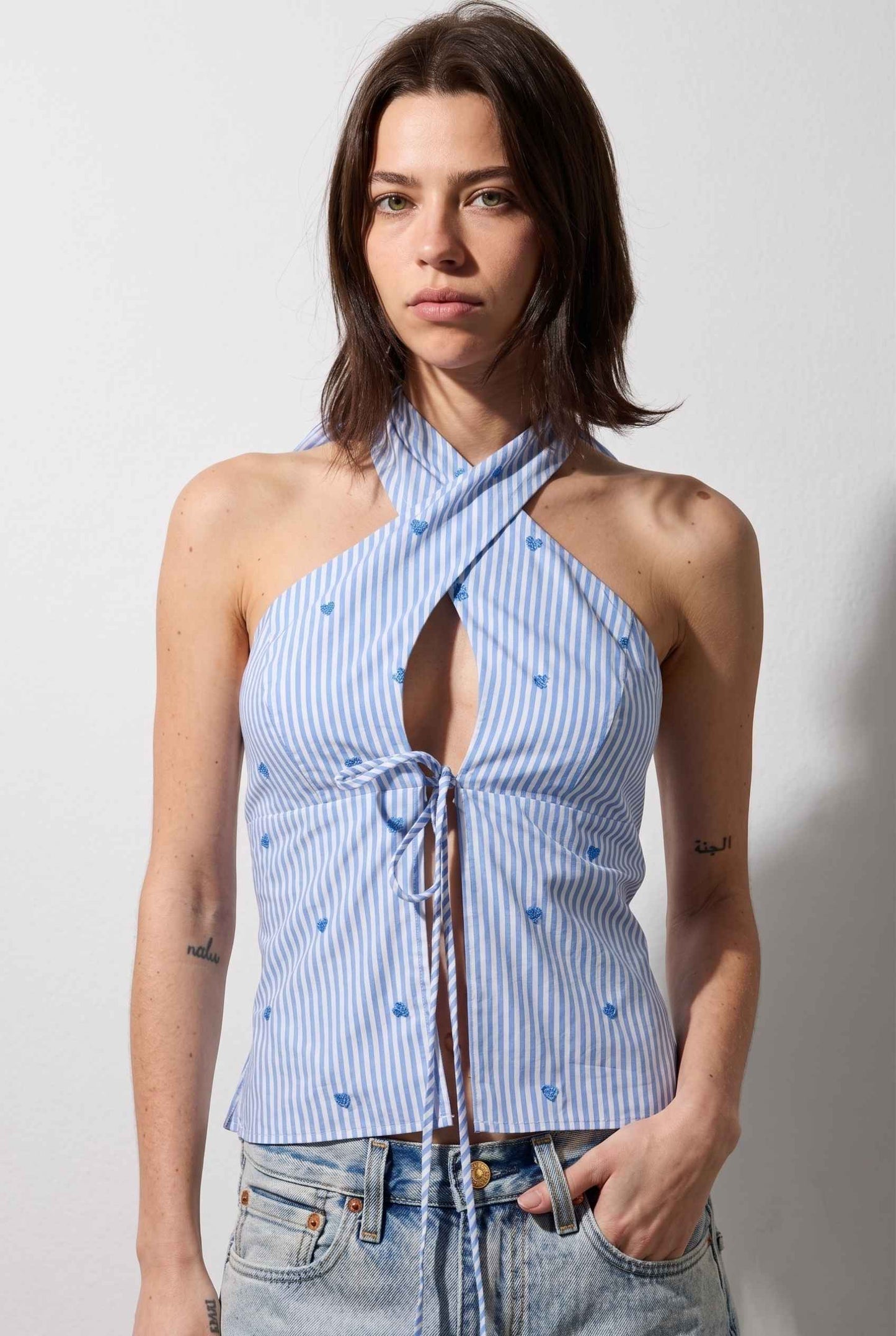 Model wearing Sara Cross-Neck Striped Cotton Halter Top Heart Embroidered Tank Top in Blue with jeans close up front tie detail