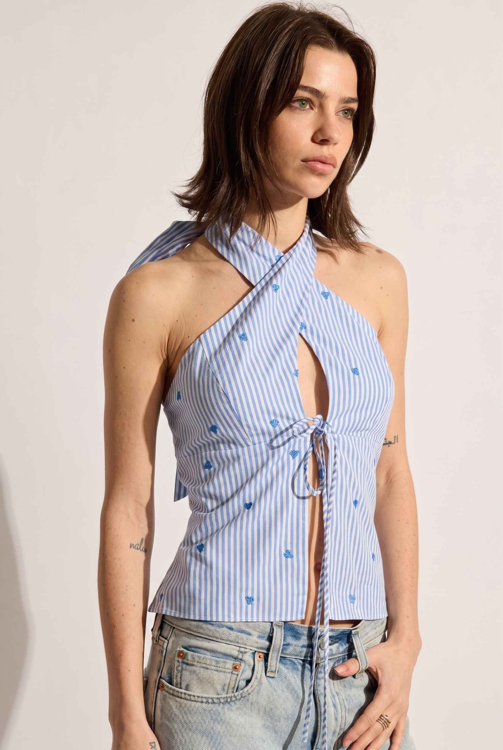 Model wearing Sara Cross-Neck Striped Cotton Halter Top Heart Embroidered Tank Top in Blue with jeans close up front tie detail
