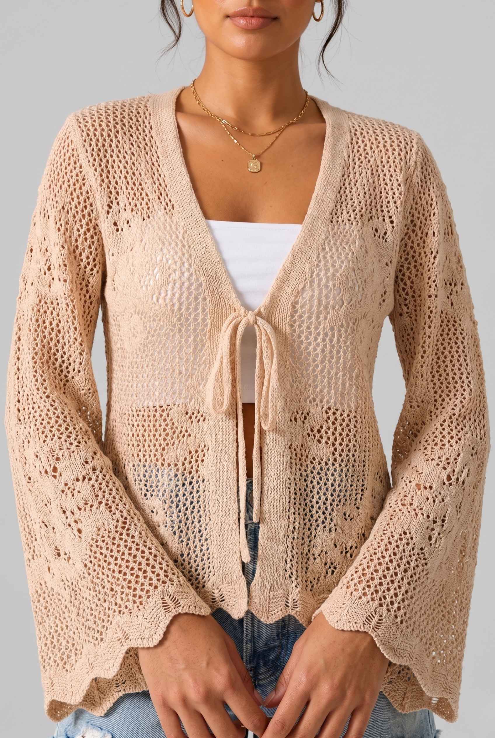 The Faye Tie-Front Cardigan Open Stitch Beige Crochet Cardigan worn by a woman on a gray background
