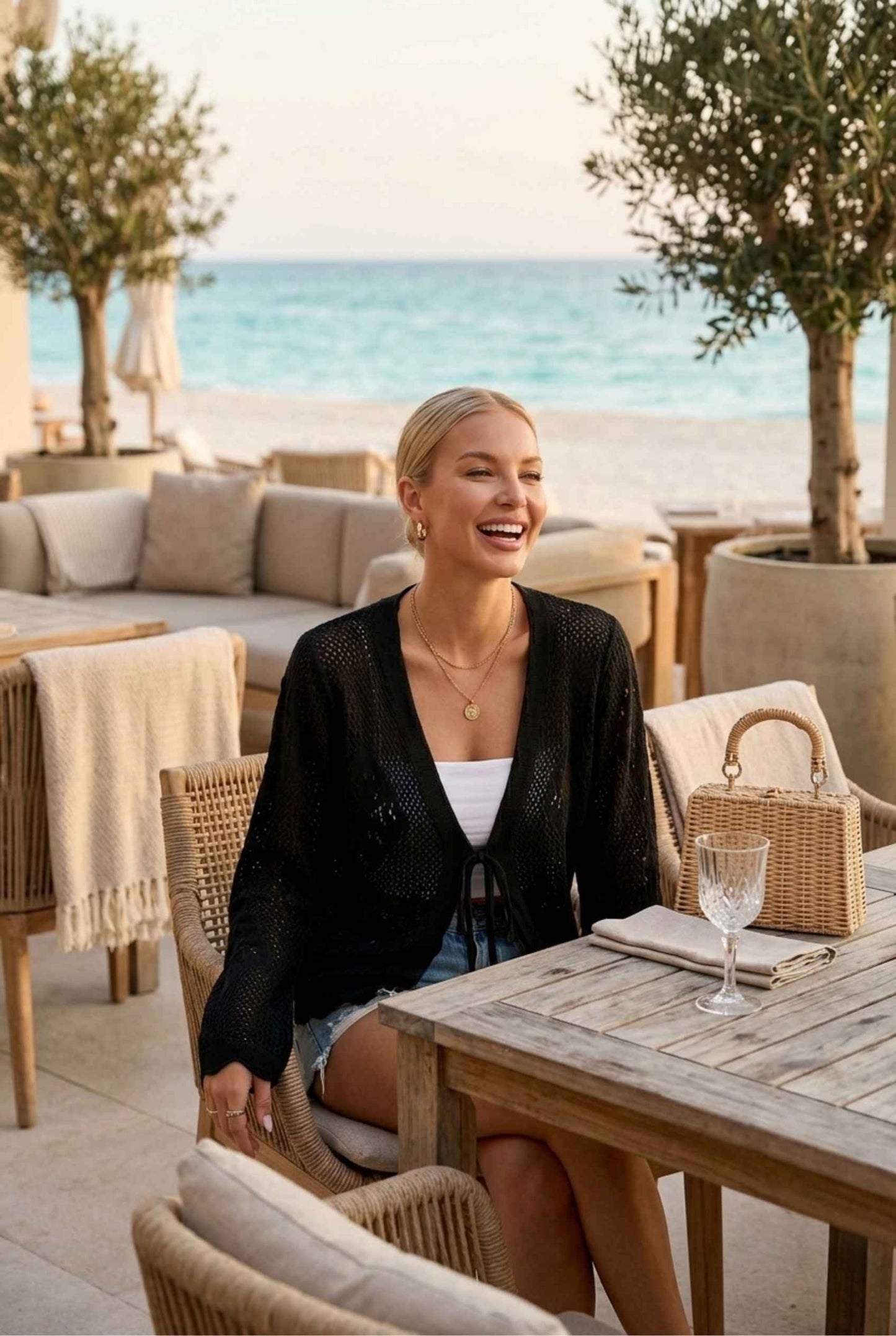 Woman wearing The Faye Tie-Front Cardigan Open Stitch Black Crochet Cardigan with denim shorts sitting at a beach restaurant smiling