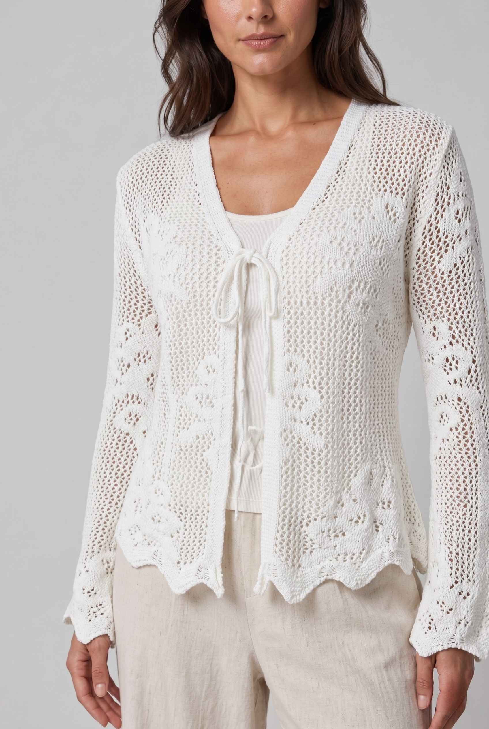 Woman wearing The Faye Tie-Front Cardigan Open Stitch White Crochet Cardigan against a gray background