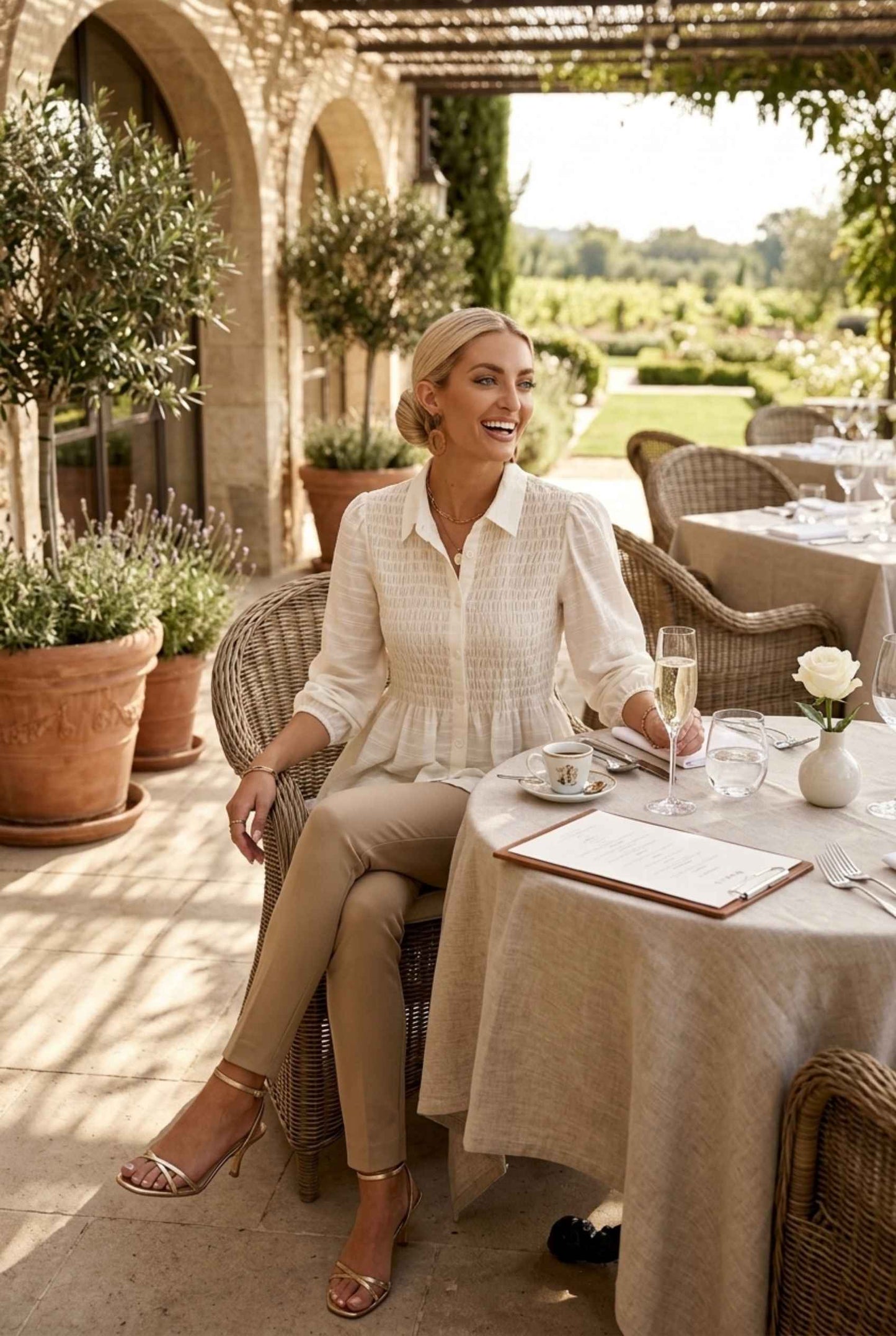 Woman wearing The Sutton Peplum Blouse Smocked Button-Down Off White Top with skinny pants and strappy sandals sitting on luxury brunch patio with a garden in the back
