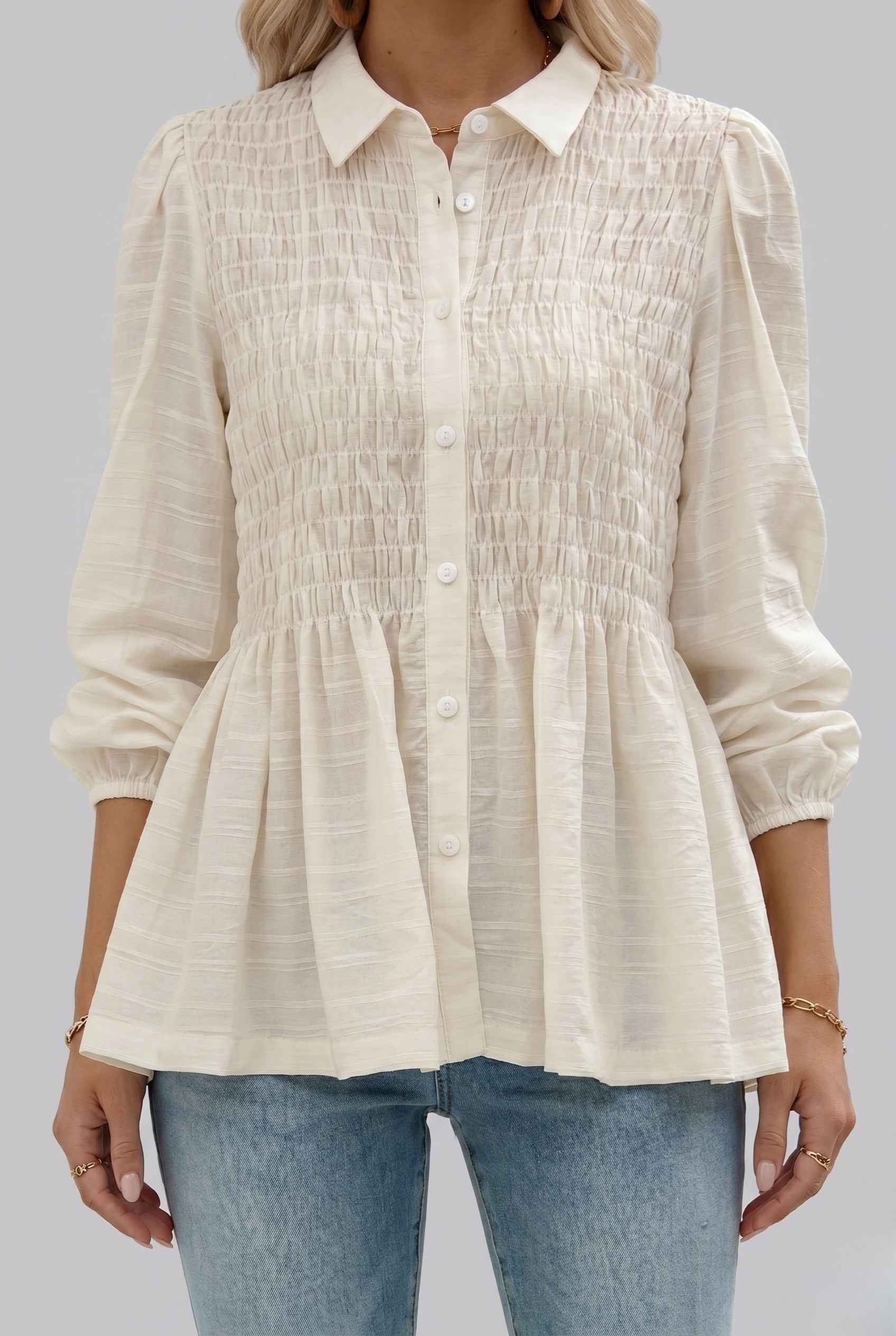 The Sutton Peplum Blouse Smocked Button-Down Off White Top worn by a woman on a gray background